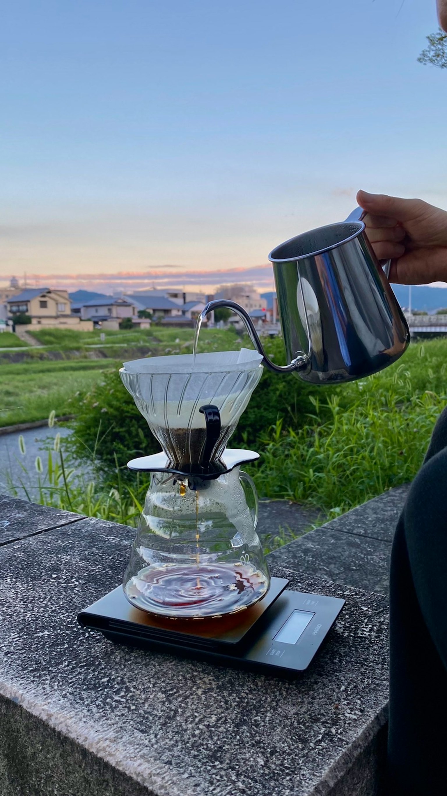 川の近くでコーヒーを淹れている様子 鋼のケトルでお湯を注ぐ手