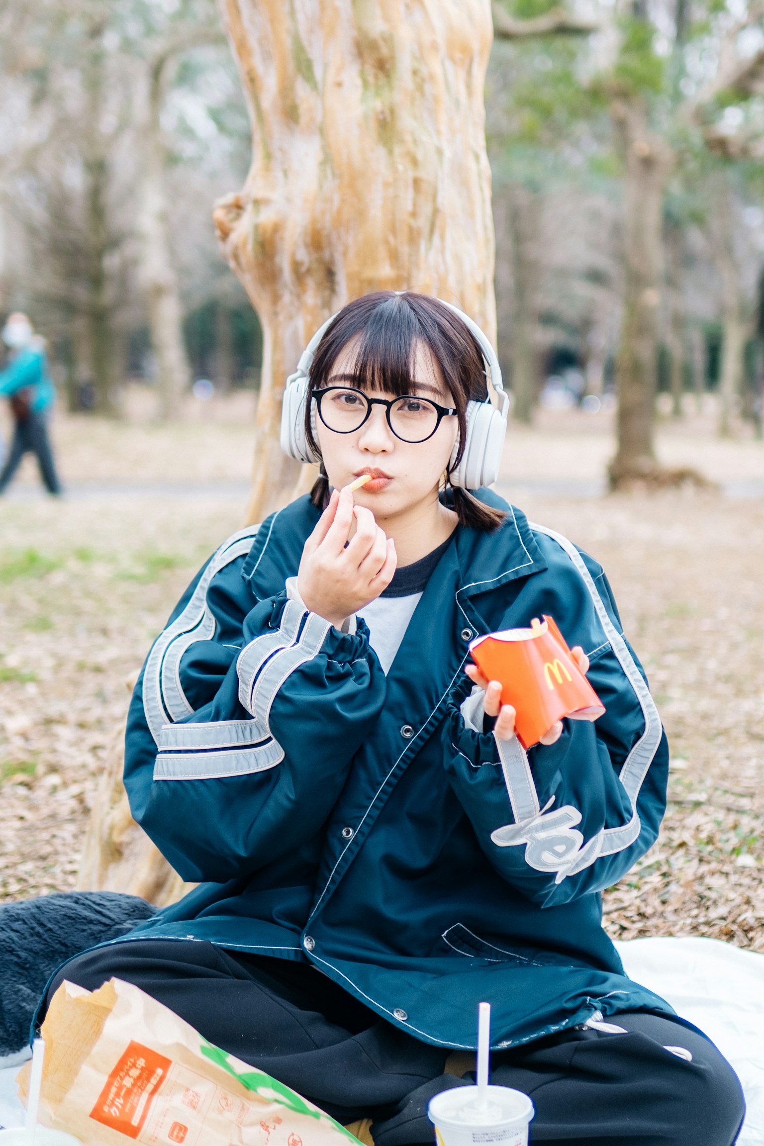 Mujer comiendo bocadillos en un parque usando auriculares y gafas vestida con pantalones negros y chaqueta azul