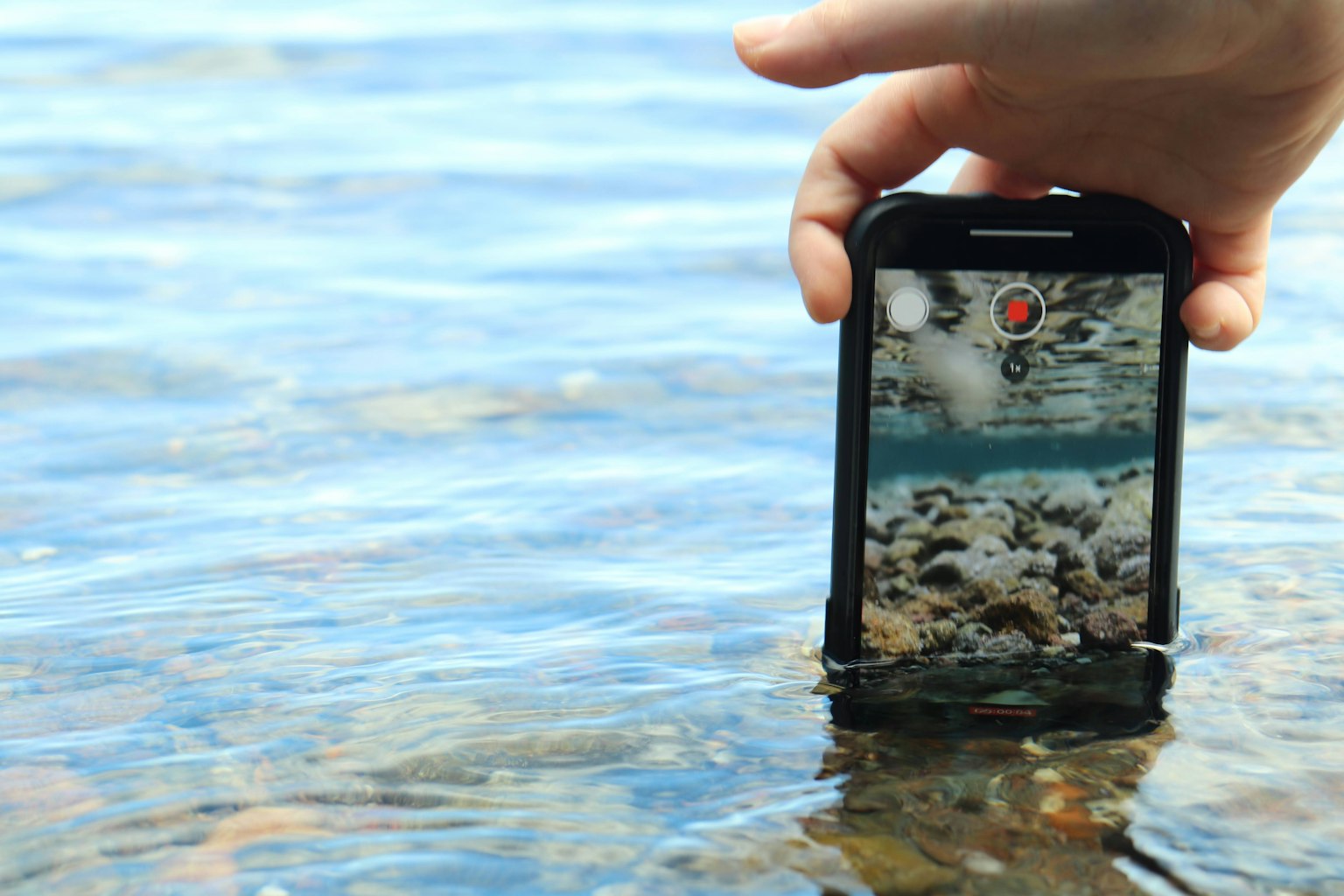 Tay cầm smartphone trên mặt nước với đá cuội nhìn thấy bên dưới