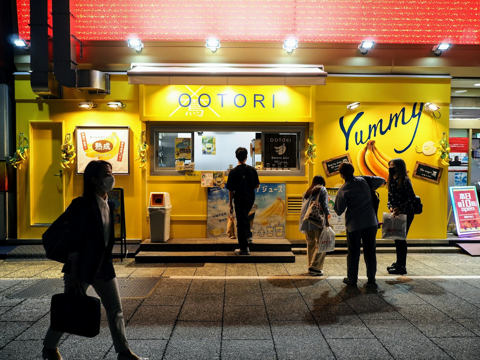 Menschen vor einem gelben Geschäft mit einem 'Yummy'-Schild und einem Menü