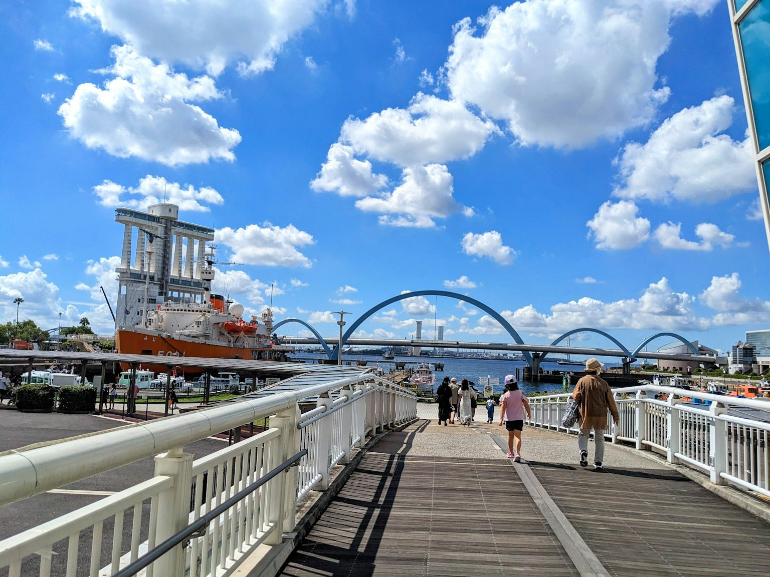 People walking on a boardwalk under a blue sky with clouds and a ship nearby