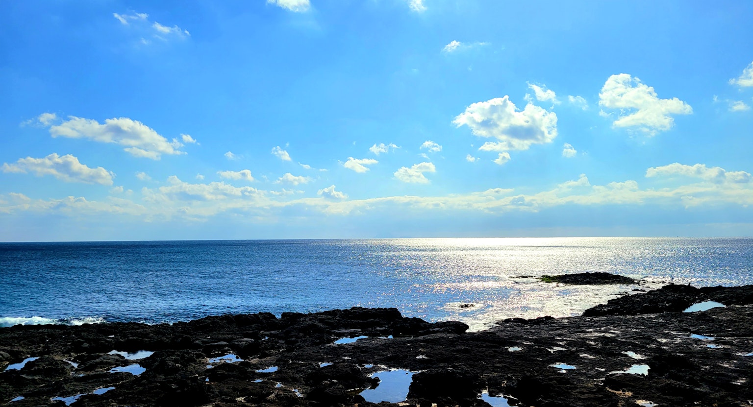 Weites blaues Meer unter einem hellen Himmel mit verstreuten Wolken und sonnenbeschienenen Felsen