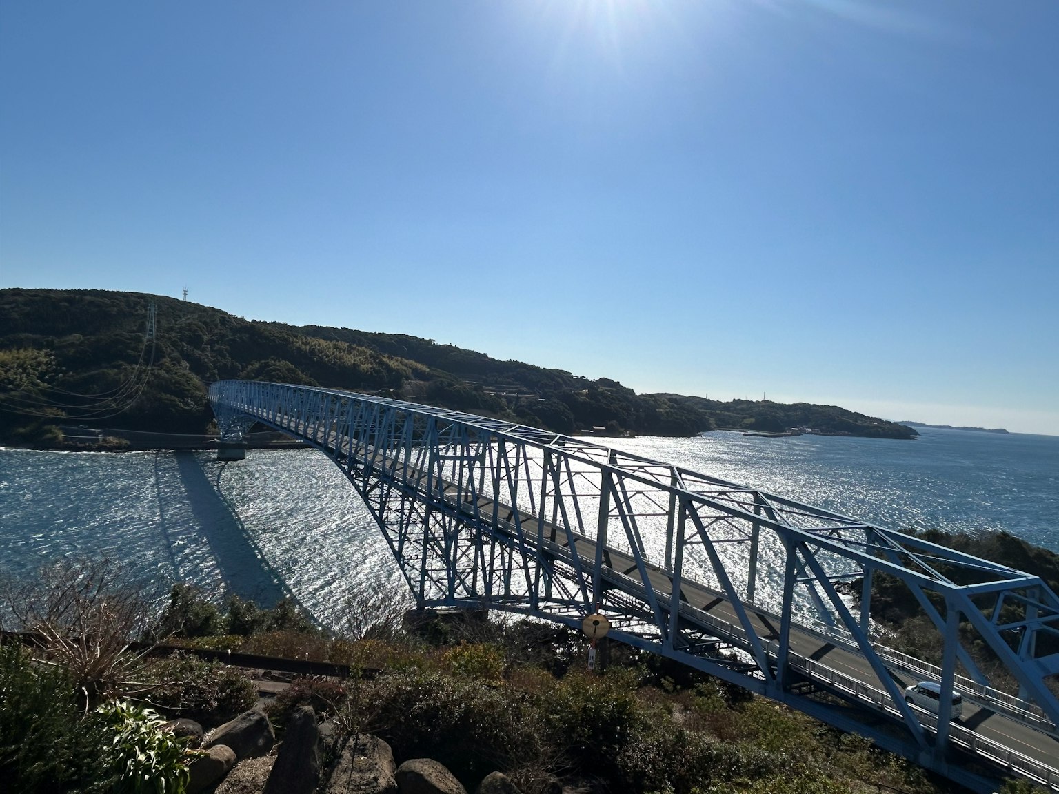 Un pont bleu s'étend au-dessus de l'eau avec des collines vertes et la lumière du soleil en arrière-plan