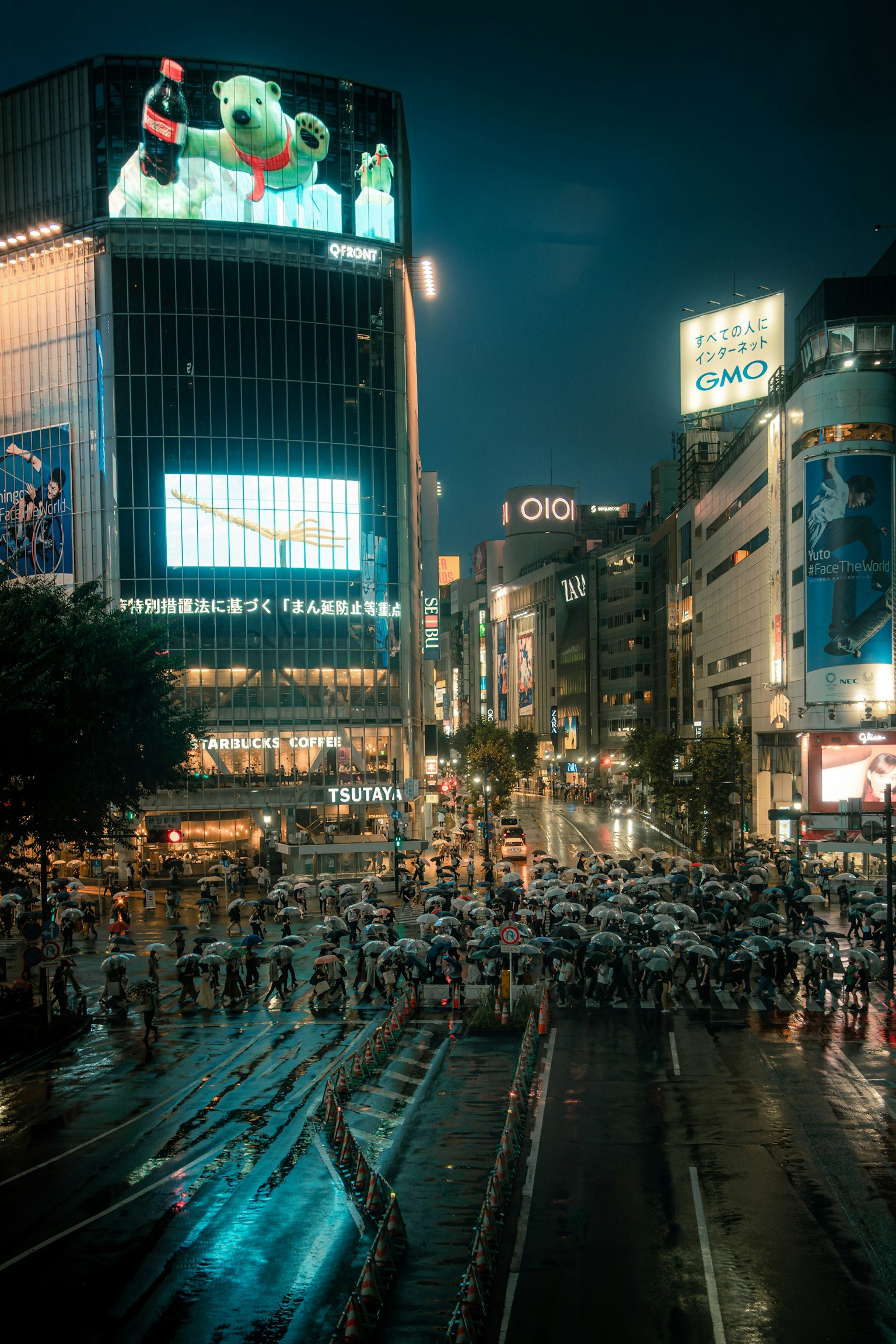 Cruce de Shibuya concurrido por la noche con una pantalla grande mostrando un personaje y personas con paraguas