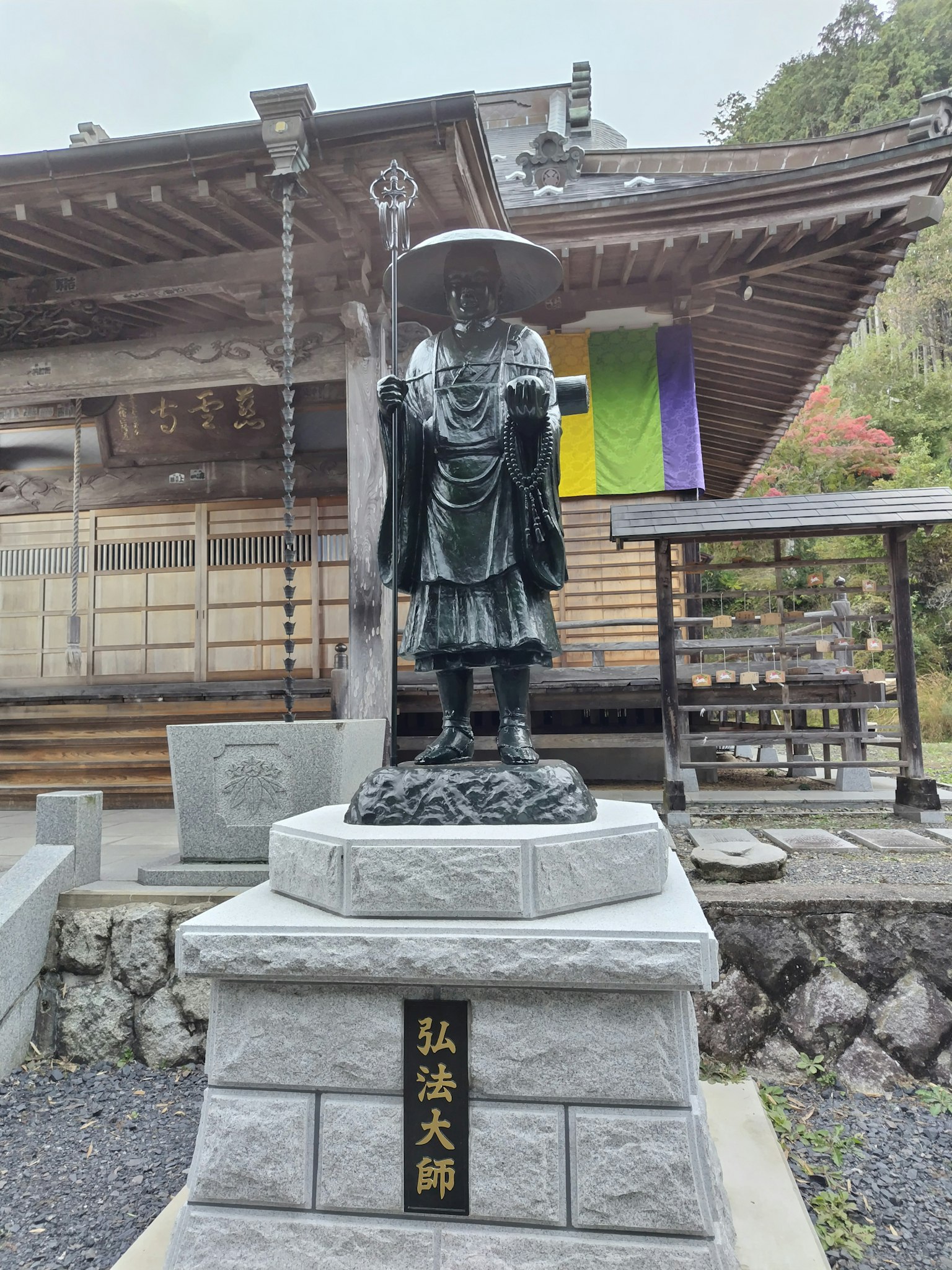 Patung Kobo Daishi di depan kuil arsitektur kayu tradisional dan dedaunan hijau