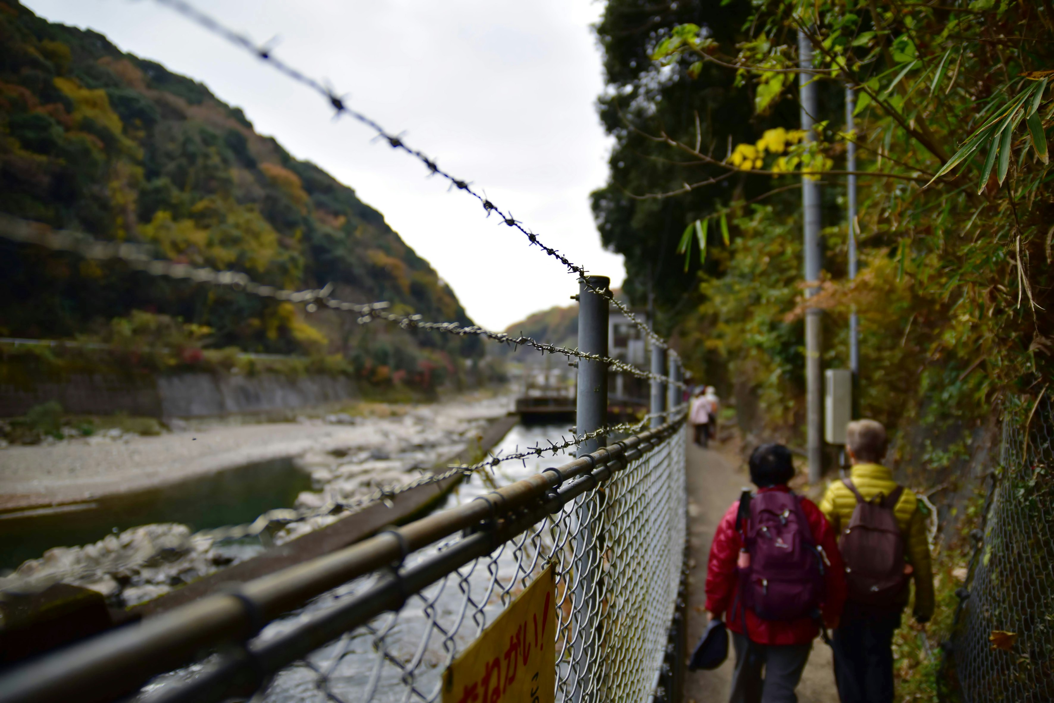 有刺鉄線のフェンス沿いに川辺を歩く人物たち（古虎渓駅付近）