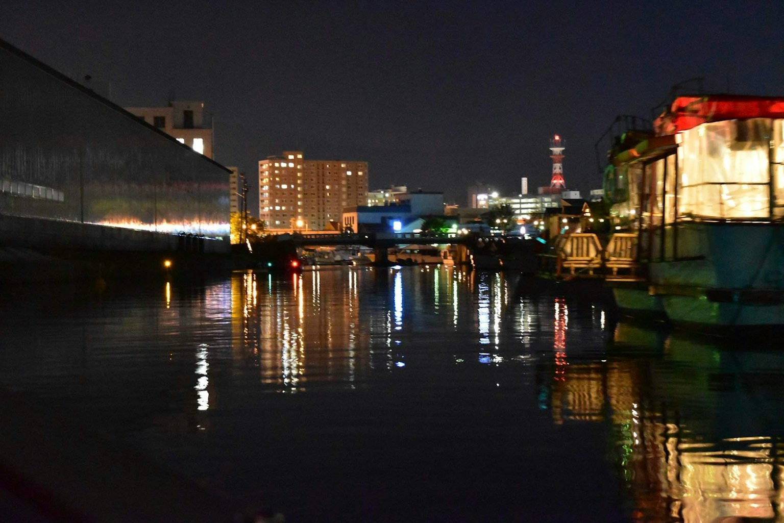 Nächtliche Flusslandschaft mit Hochhäusern und reflektierten Lichtern sowie festgemachten Booten