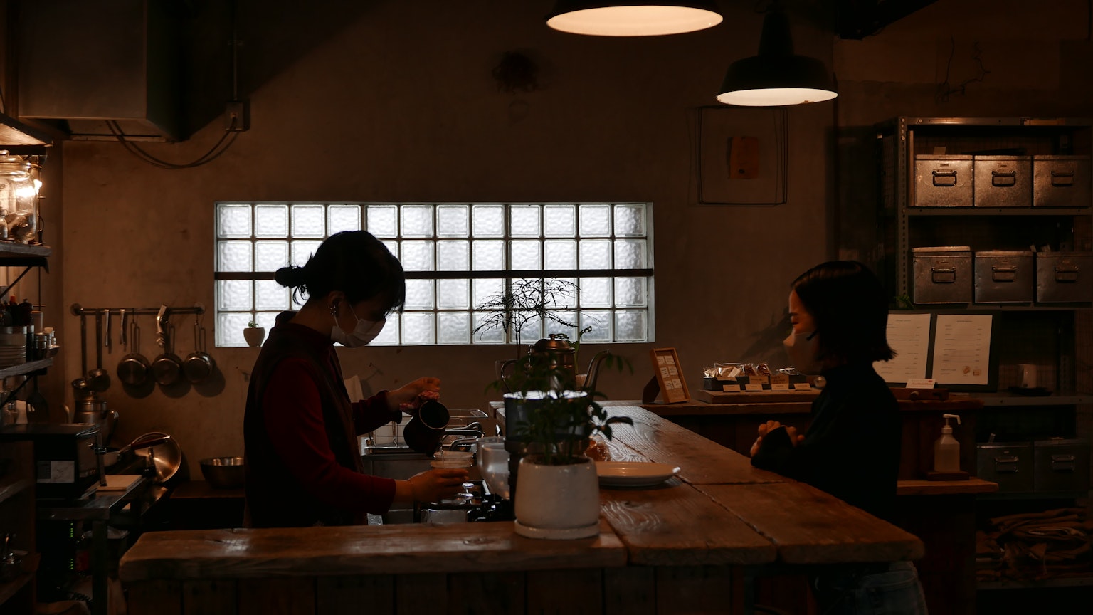 Adegan interior kafe dengan seorang wanita yang bekerja dan satu wanita yang duduk Pencahayaan lembut menciptakan suasana hangat