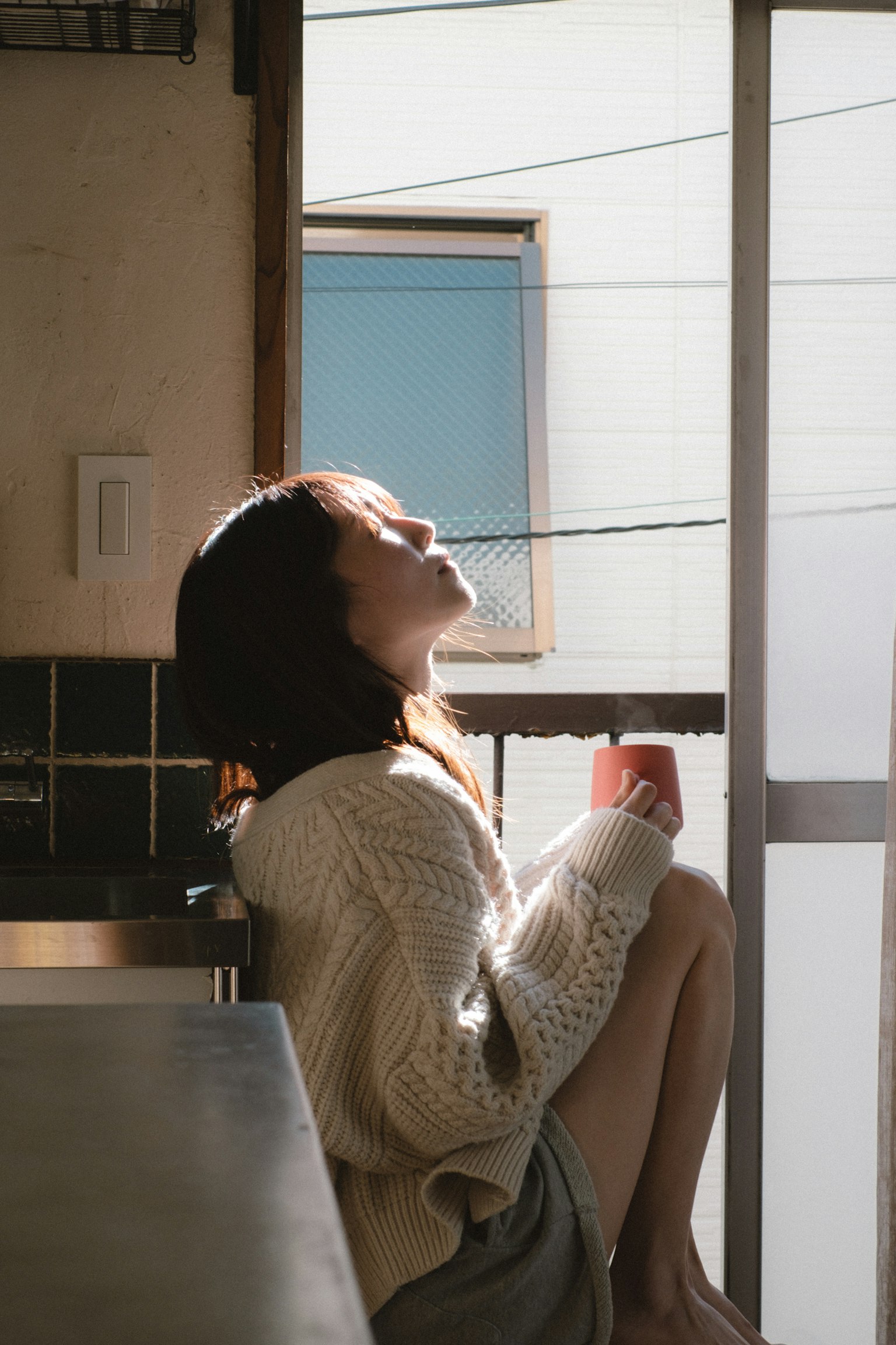 Frau sitzt am Fenster mit einer Tasse Kaffee und genießt das Sonnenlicht