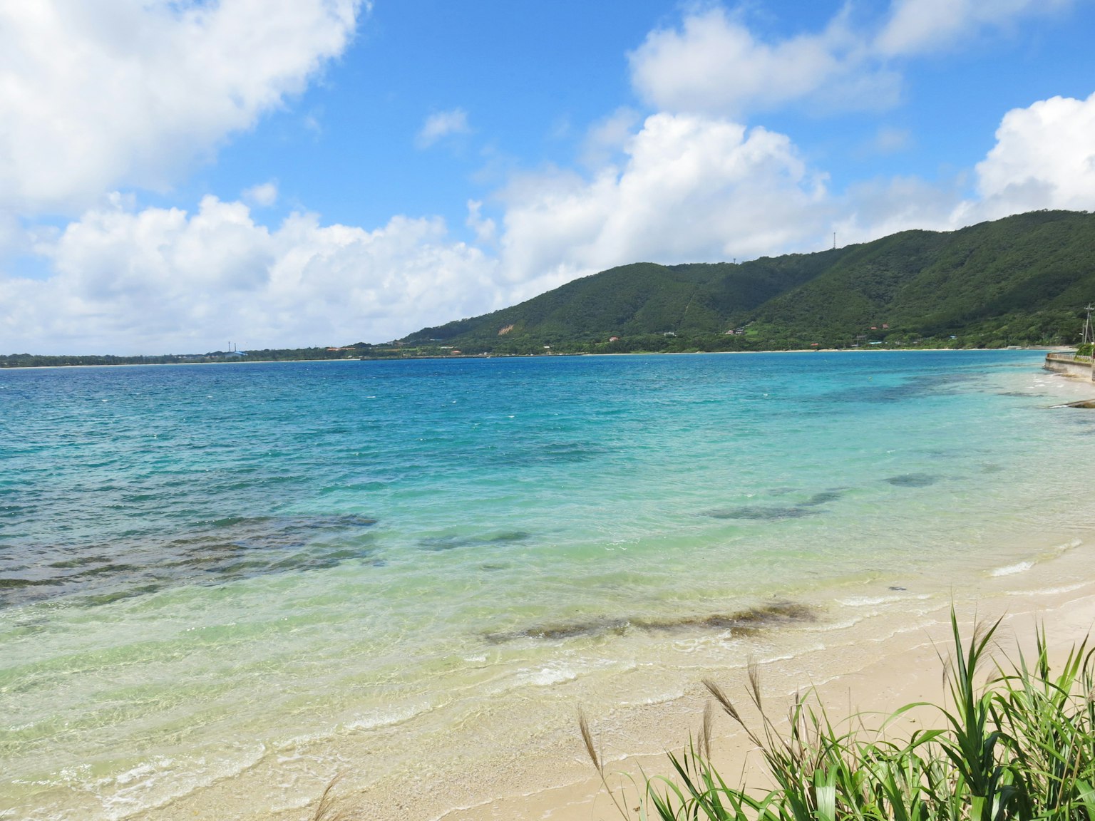 Scenic view of blue sea and white sandy beach with green hills and blue sky