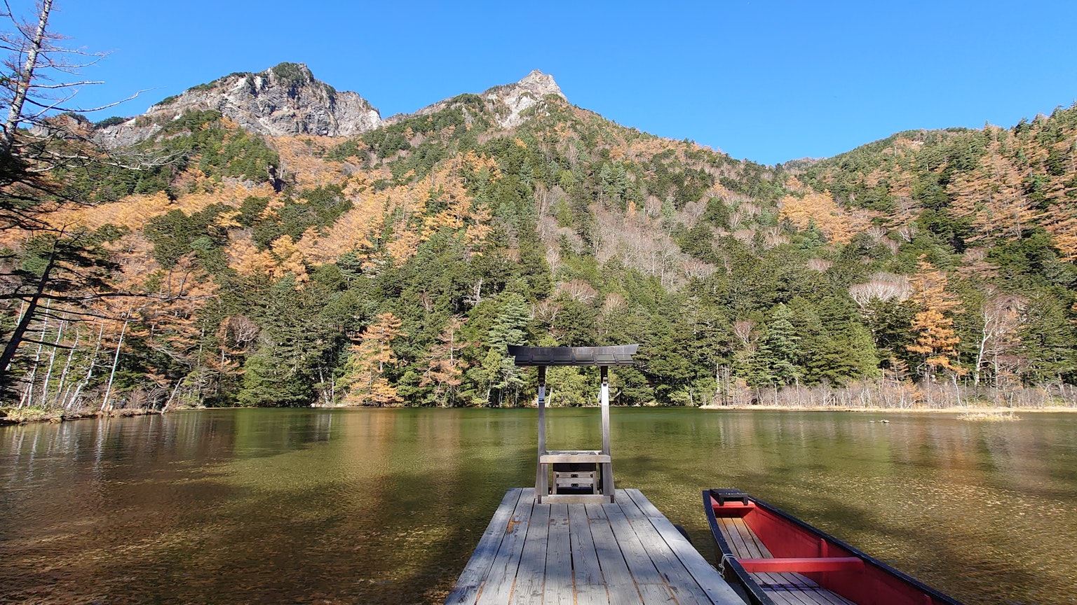 Malerscher Blick auf einen ruhigen See und Berge mit einem Steg im Vordergrund, der Herbstfarben zeigt