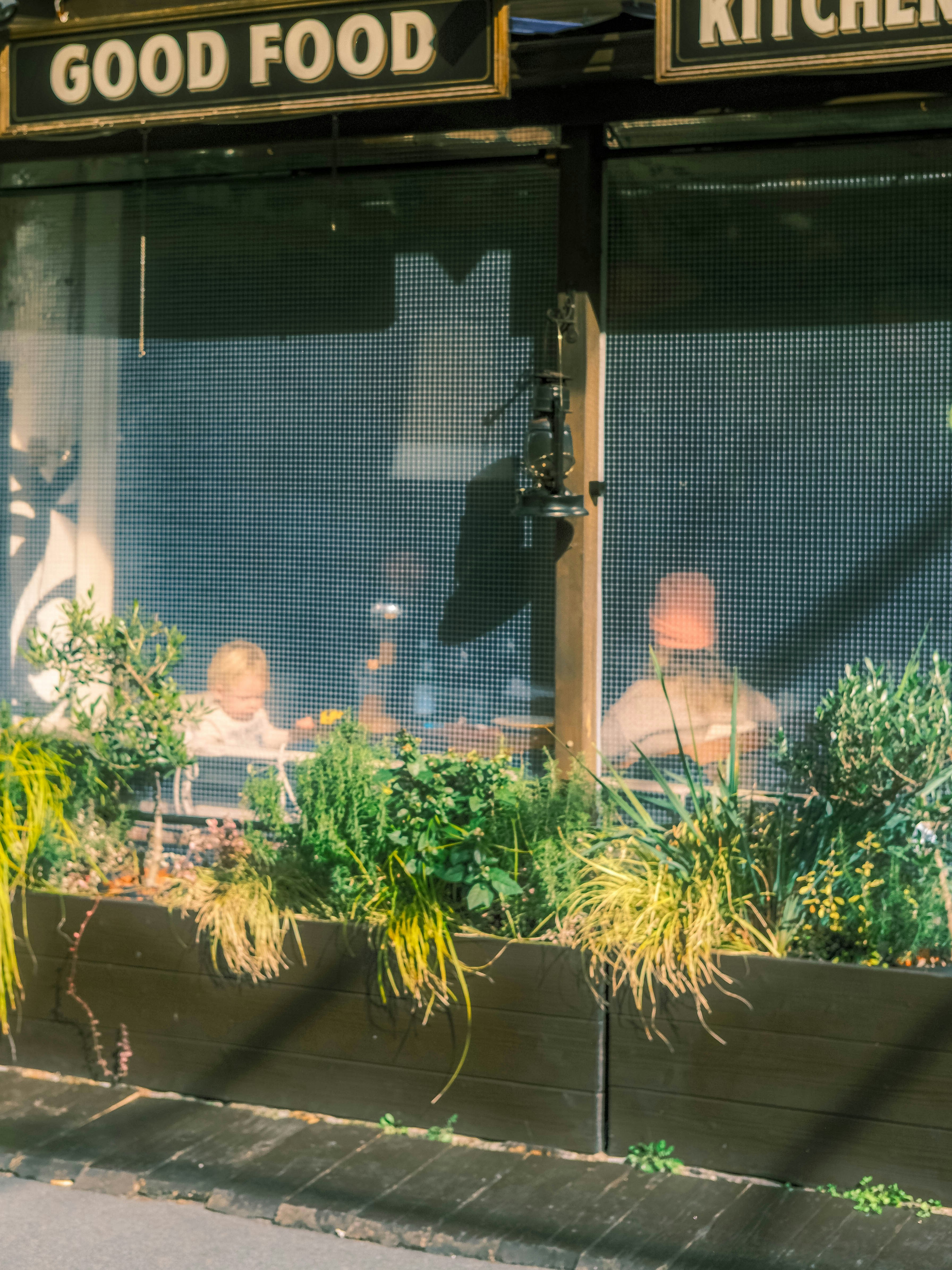 people seen through planters in front of a storefront with a “GOOD FOOD” sign
