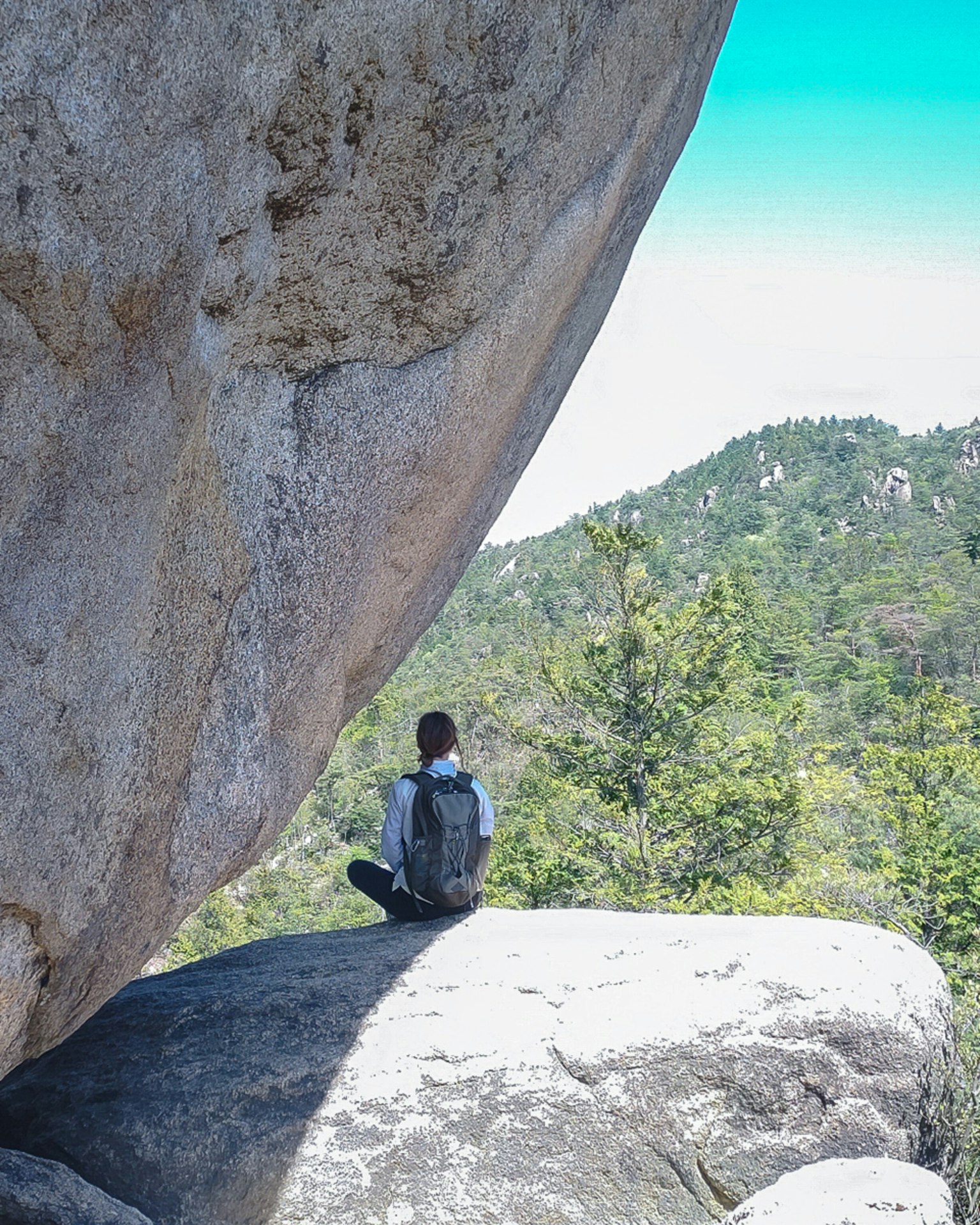 Person sitting near a large rock with green mountains in the background