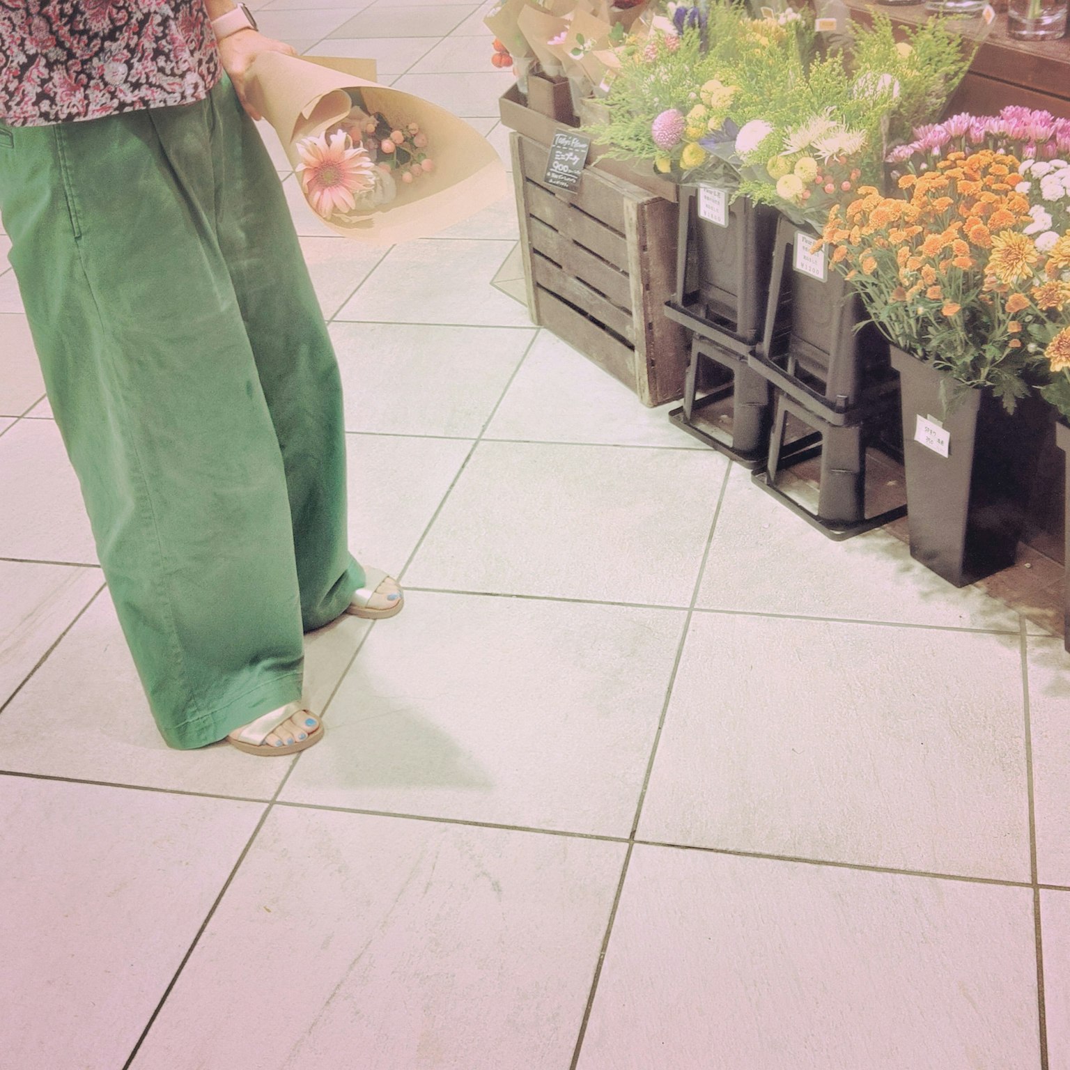 A woman wearing green pants holding a bouquet stands in front of a flower shop