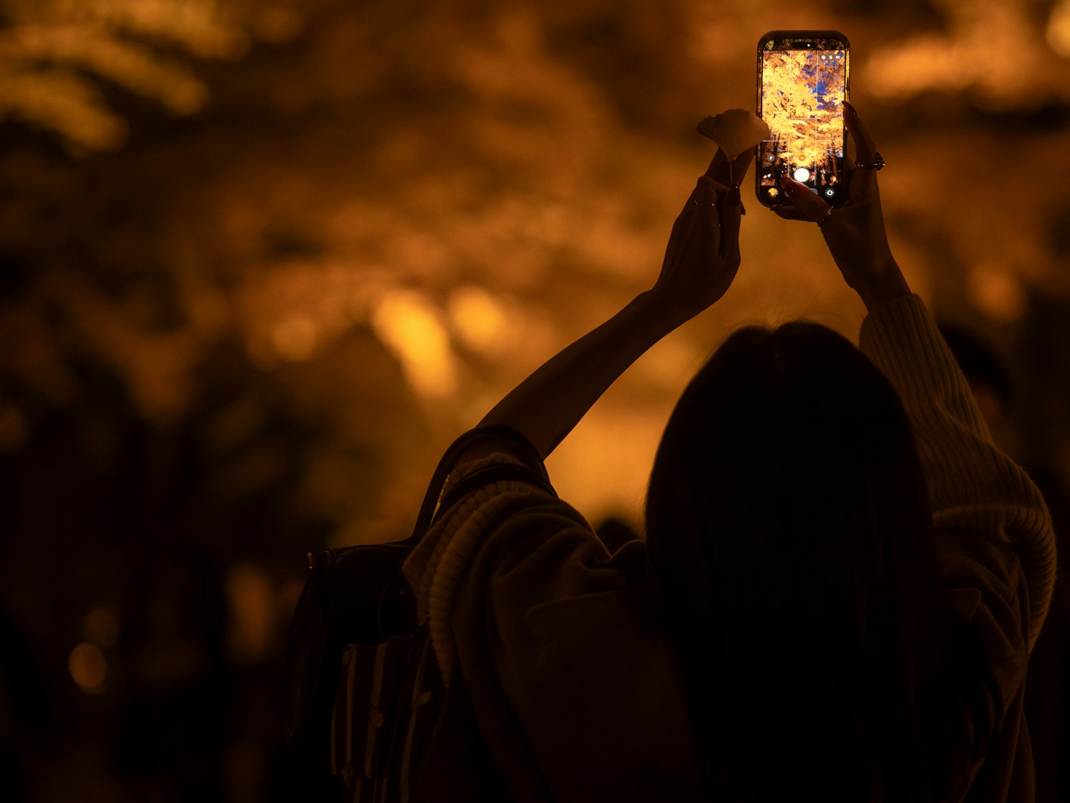 女性がスマートフォンで写真を撮っている夜のシーン 温かいオレンジ色の照明が周囲を照らしている