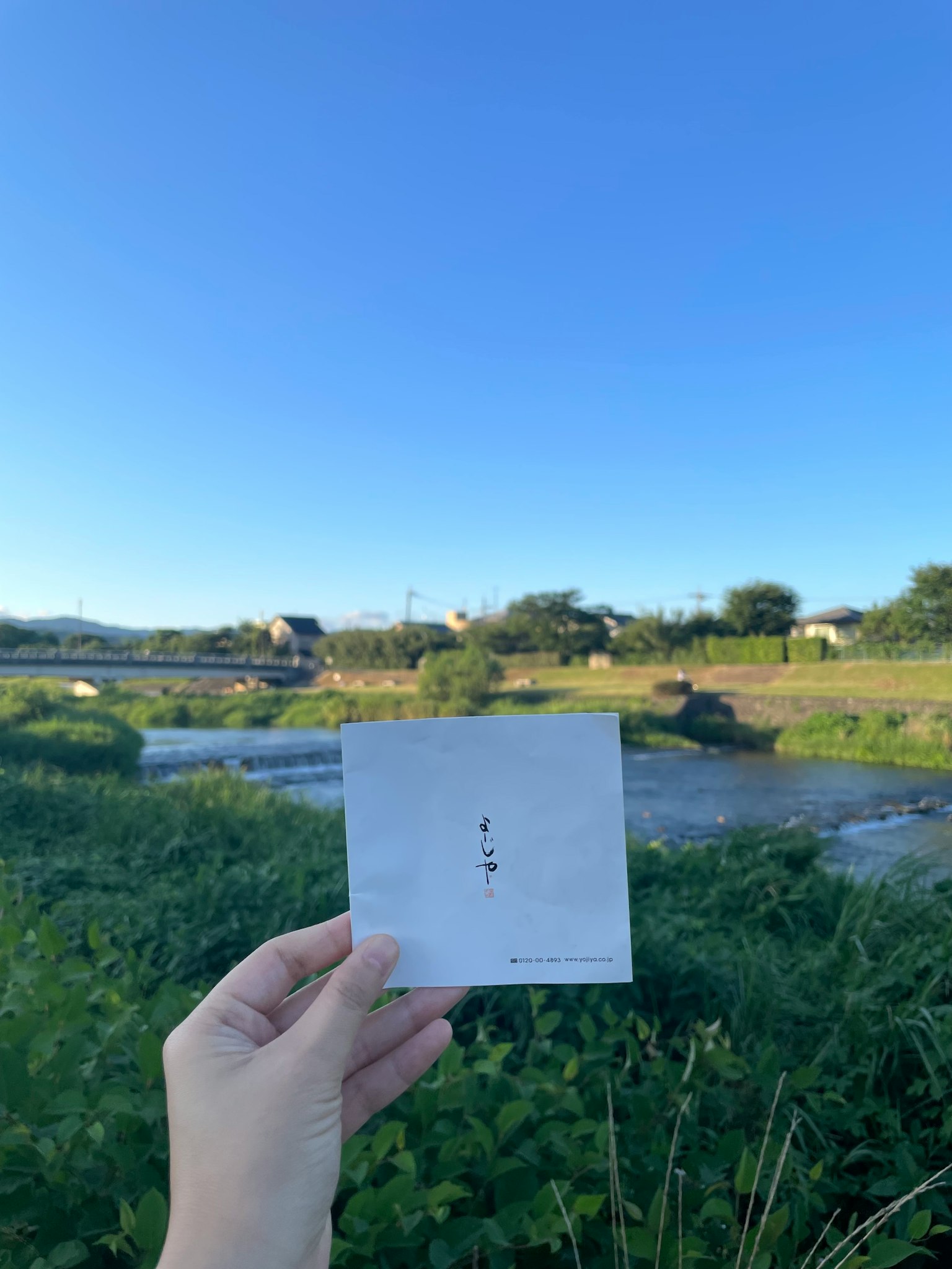 一隻手拿著白紙，背景是晴朗的藍天和河流風景