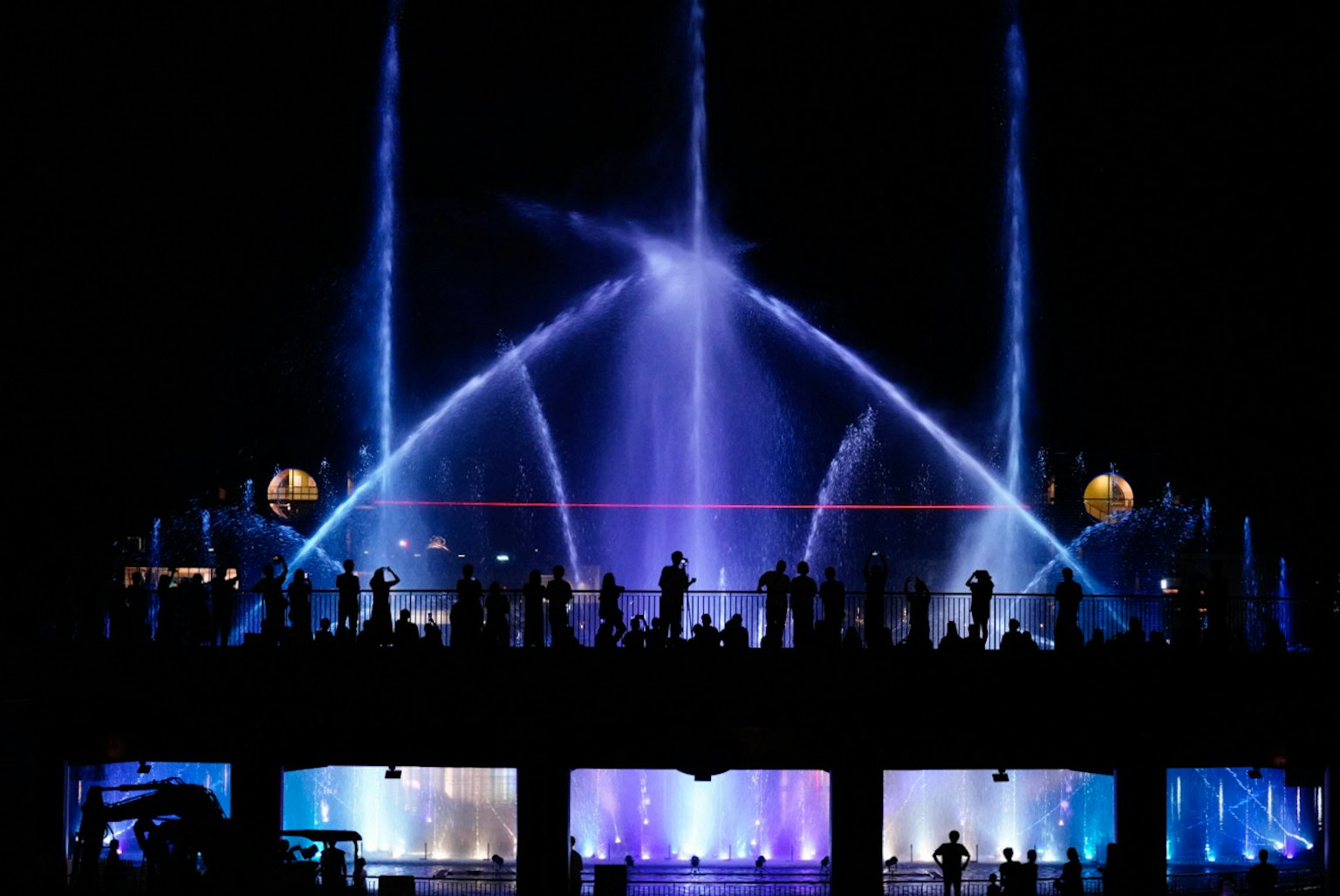 การแสดงน้ำพุในเวลากลางคืนพร้อมแสงสีน้ำเงินสดใสและผู้ชมในเงา