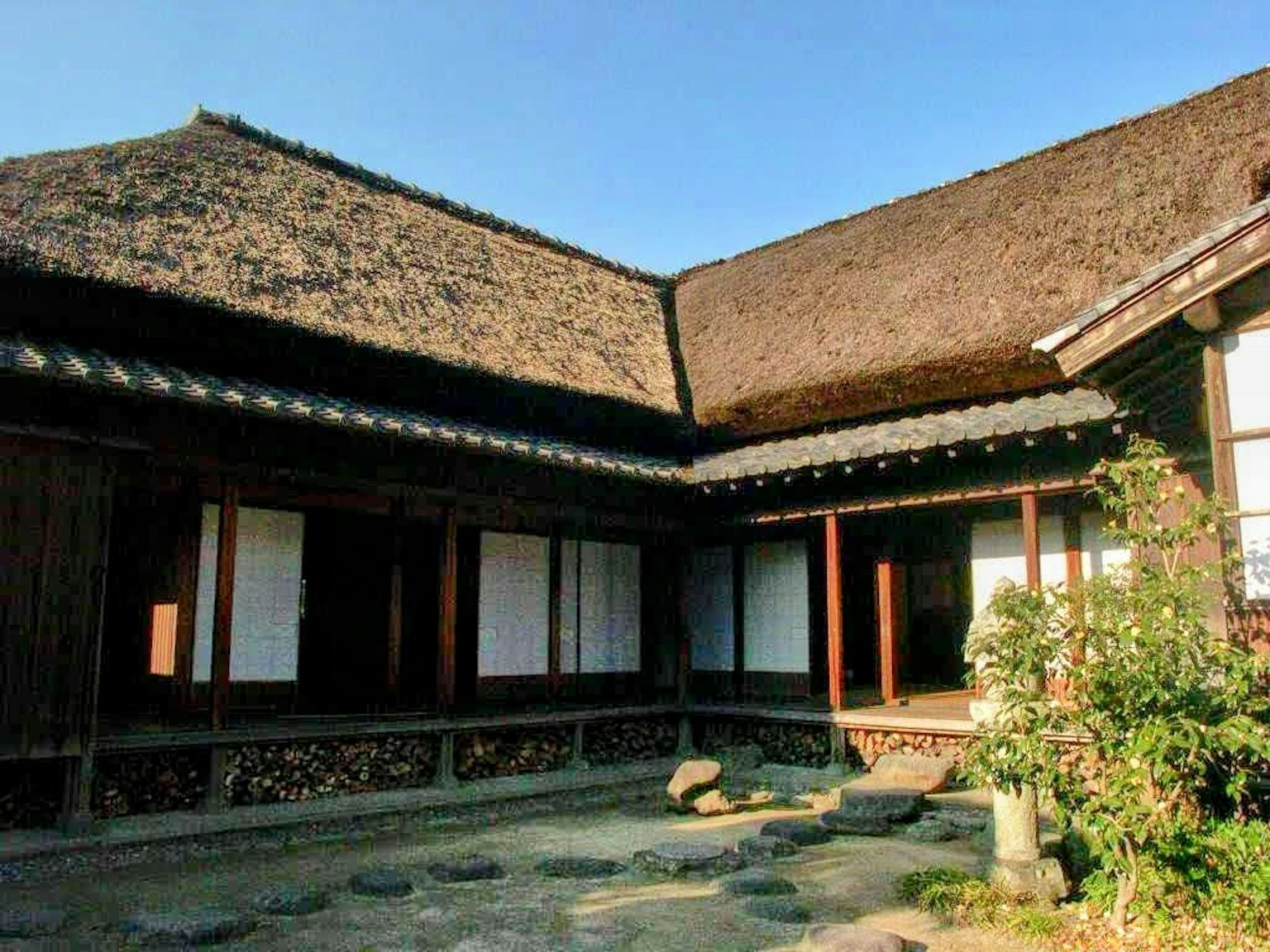 Extérieur d'une maison japonaise traditionnelle avec un toit en chaume et une structure en bois, avec un chemin en pierre et des plantes dans le jardin