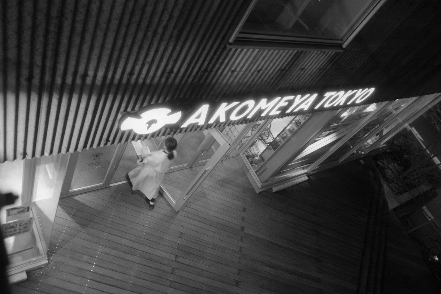 Image en noir et blanc présentant le panneau et la vitrine d'Akomeya Tokyo dans un cadre urbain