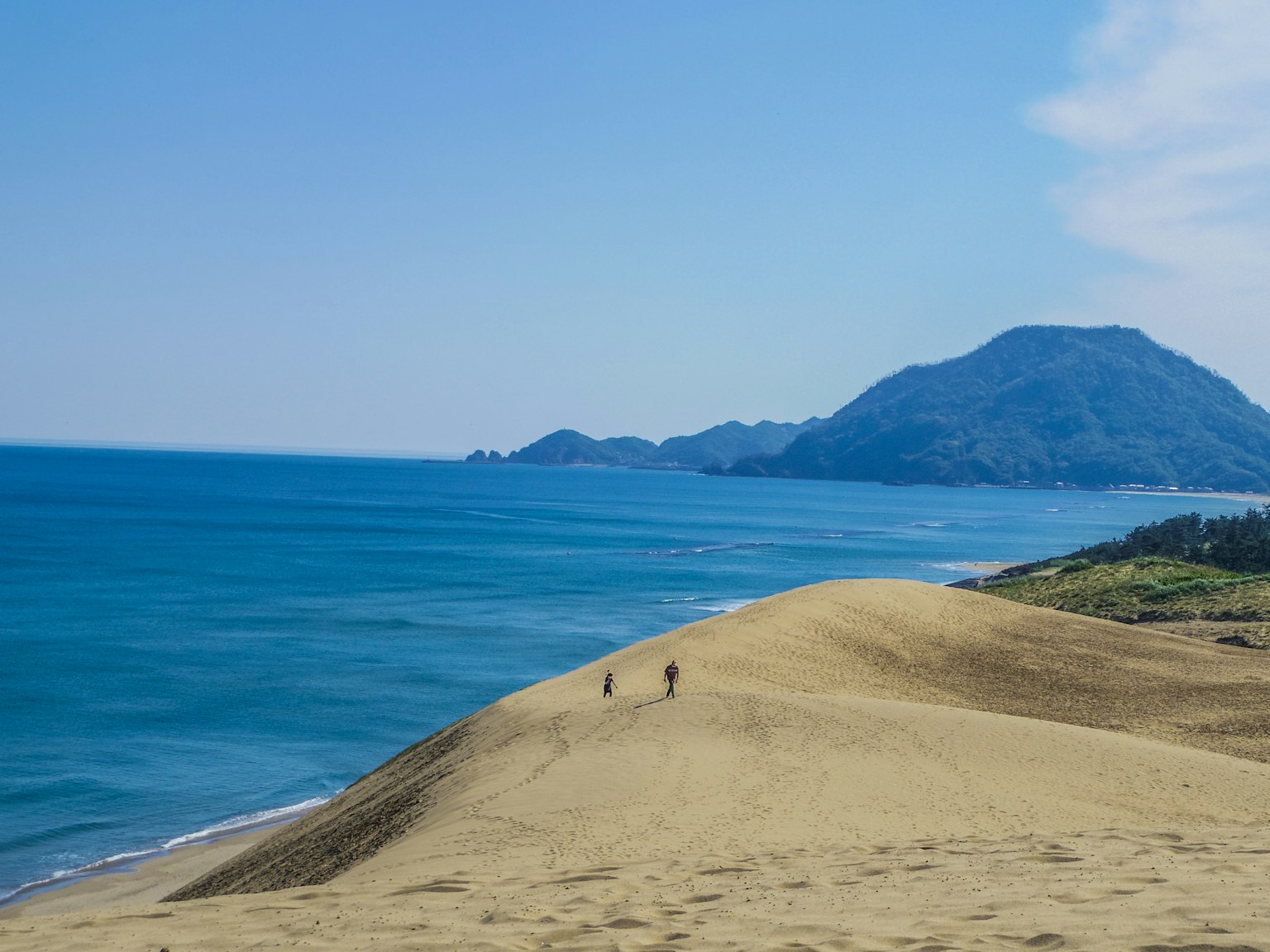 Des personnes marchant sur des dunes de sable avec un arrière-plan de mer bleue et de montagnes