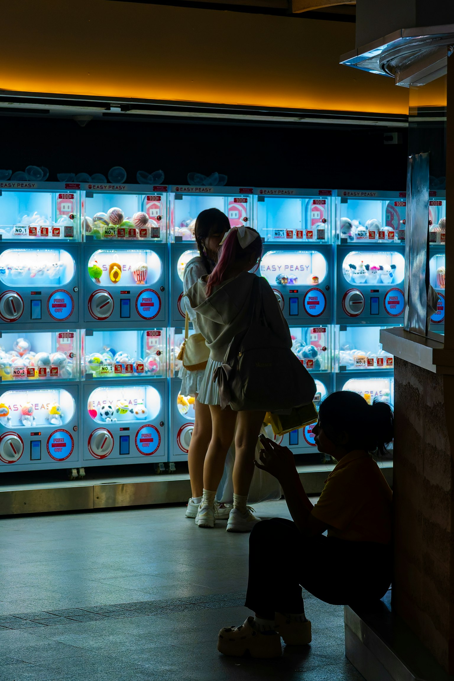 Two women embracing in front of vending machines with colorful capsules and a man sitting nearby