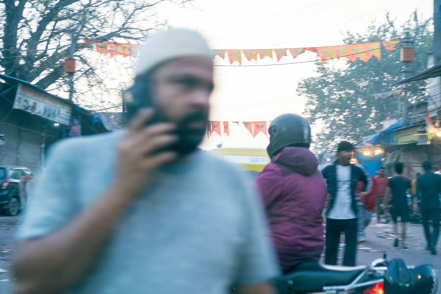 Một người đàn ông đang nói chuyện điện thoại trên đường phố ngoài trời với mọi người trên xe máy và cờ trang trí ở phía sau