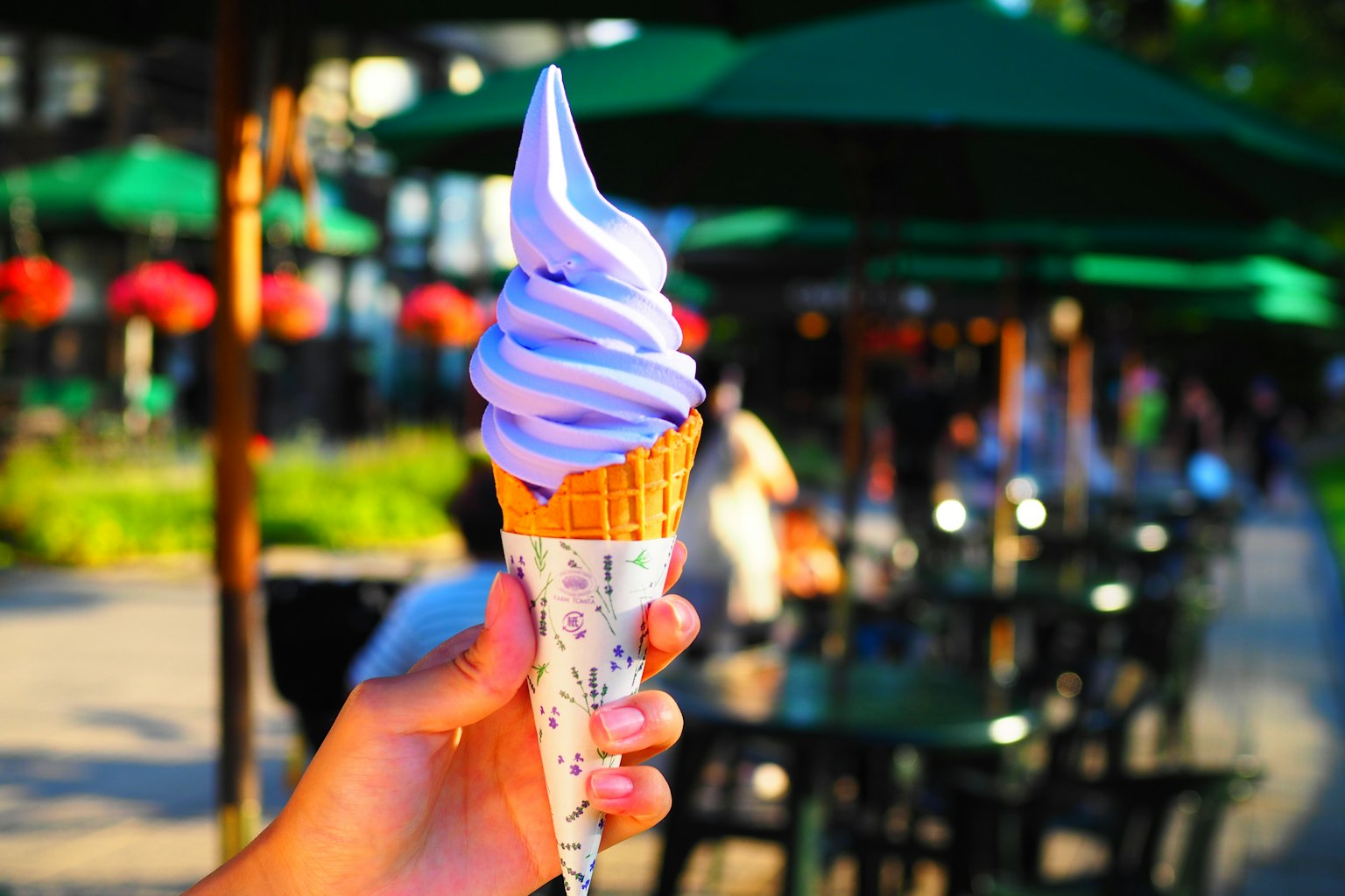 Sebuah tangan memegang cone es krim lembut ungu dengan payung hijau dan orang-orang di latar belakang