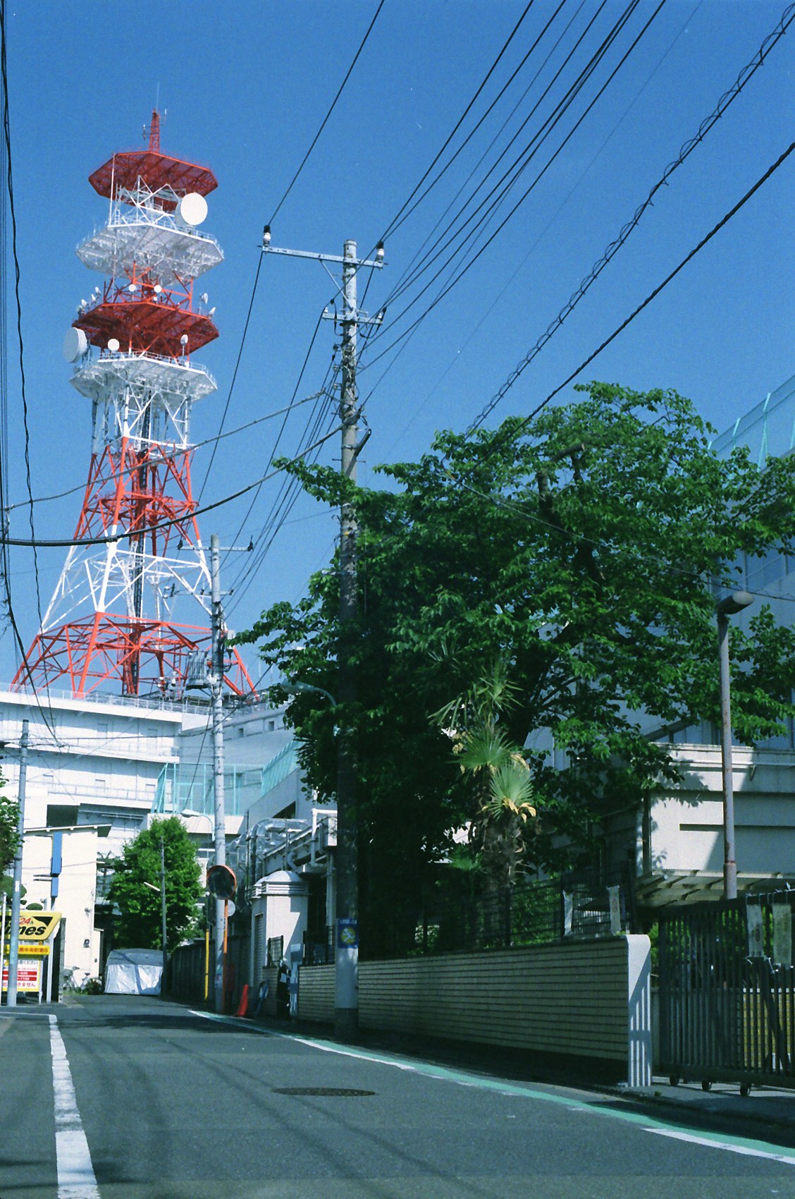 Red-and-white transmission tower beside utility poles and power lines on a street under blue sky