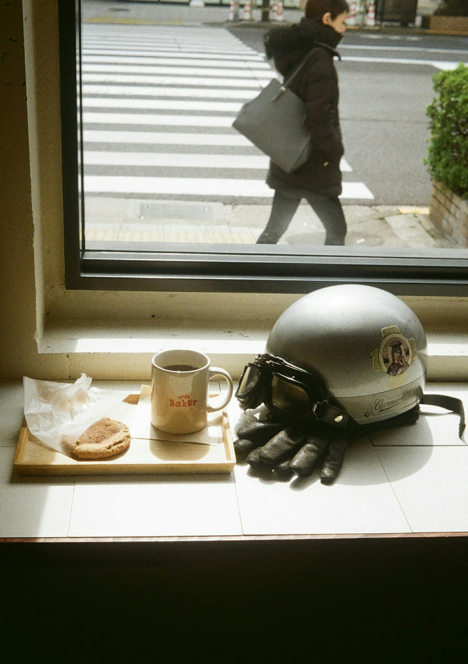 Una escena con un casco y guantes junto a una taza de café y una galleta cerca de una ventana un peatón cruzando la calle