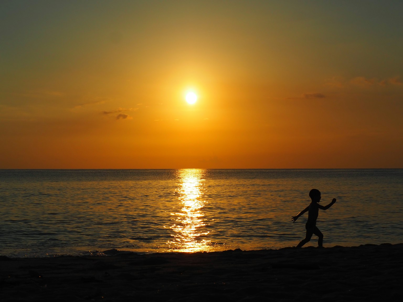 Silhouette eines Kindes, das gegen einen Sonnenuntergang über dem Ozean mit orangefarbenen Farbtönen im Himmel läuft