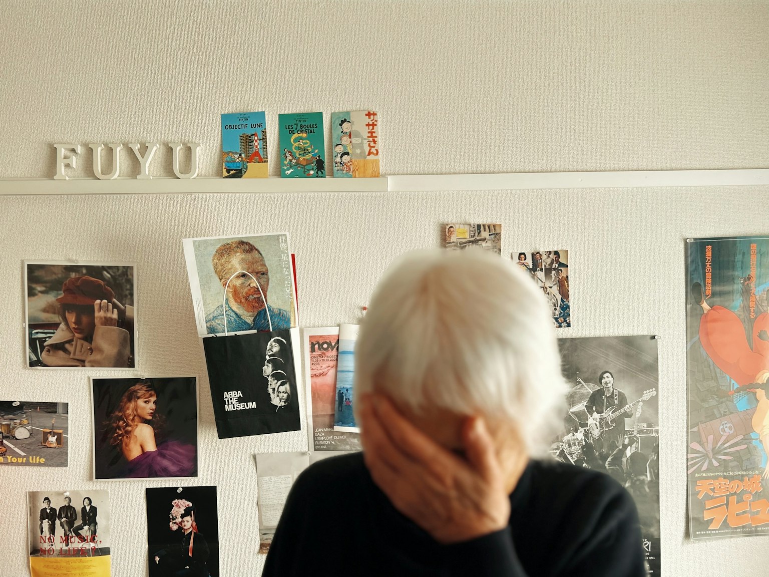 白髪の人物が顔を手で覆っている部屋の中の様子 壁には多くの写真やポスターが貼られている "FUYU" の文字が見える