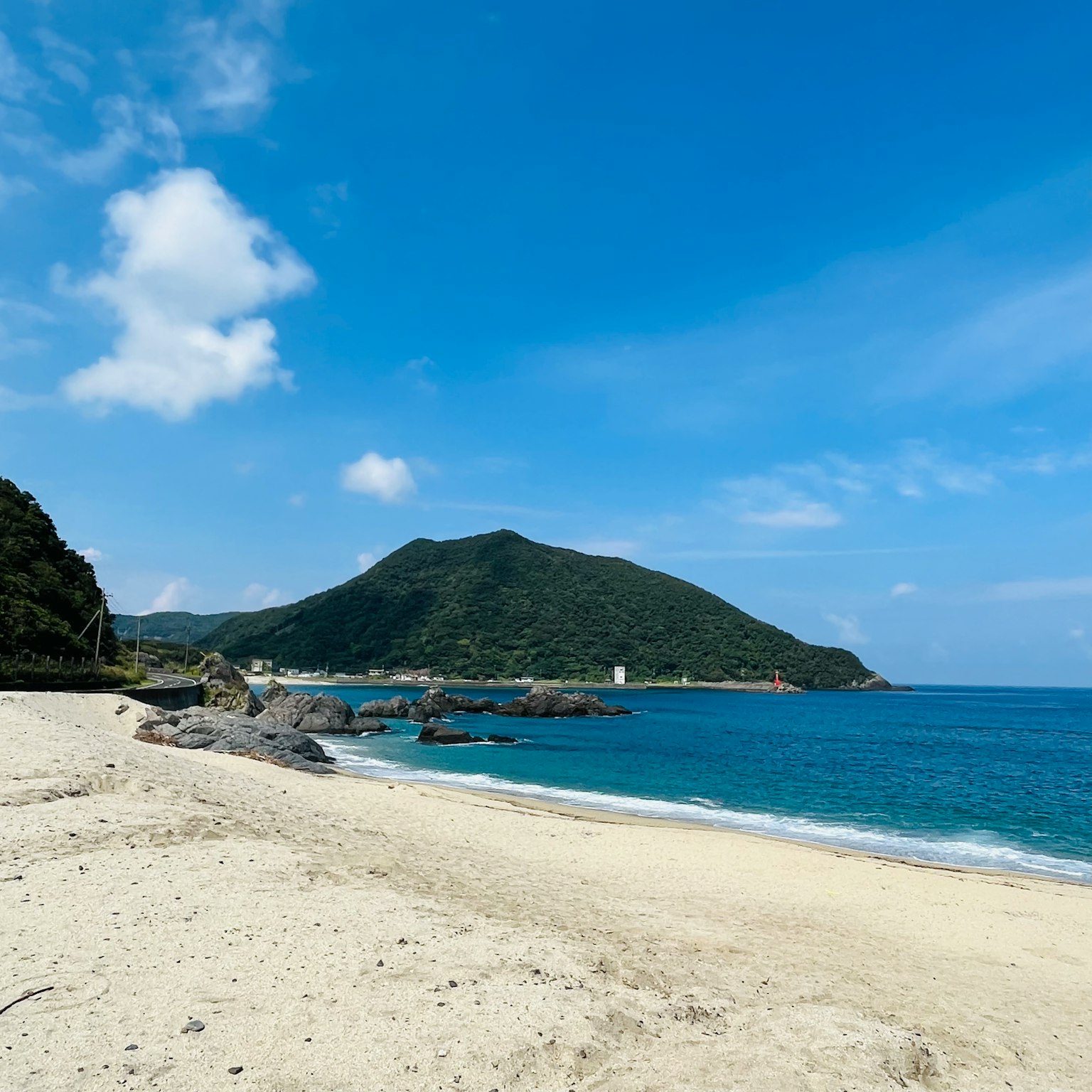 ทิวทัศน์ชายหาดที่มีทะเลสีฟ้าและทรายขาว ภูเขาสีเขียวอยู่เบื้องหลัง