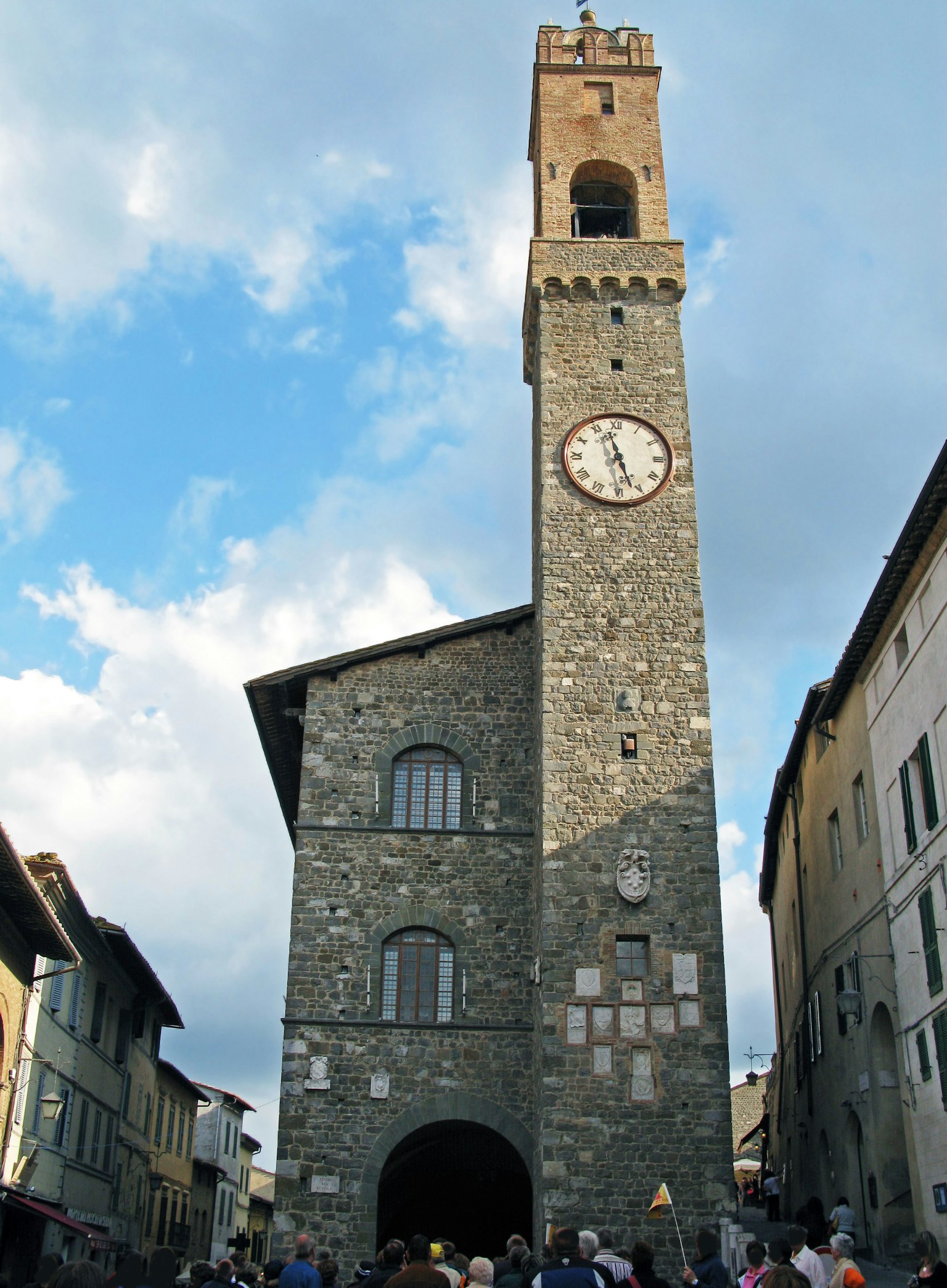 Alter Steinturm mit Uhr auf einem Platz voller Menschen unter einem blauen Himmel
