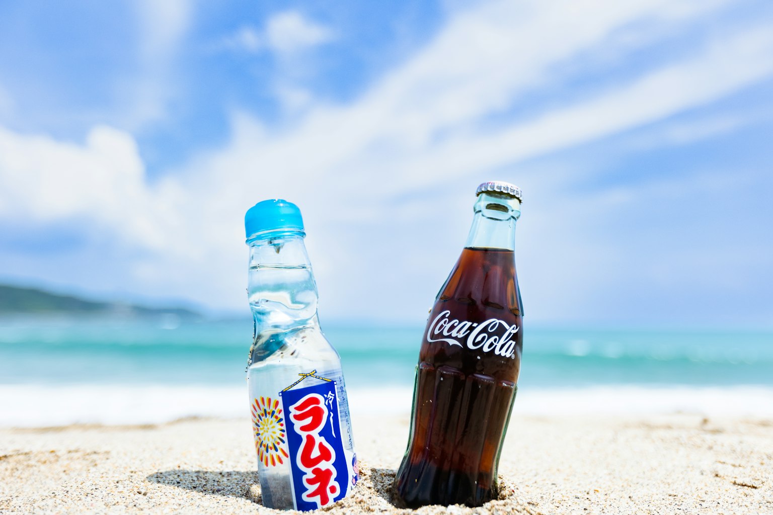 ขวดเครื่องดื่มสองขวดบนชายหาด Ramune และ Coca-Cola มีท้องฟ้าสีฟ้าและมหาสมุทรเป็นพื้นหลัง