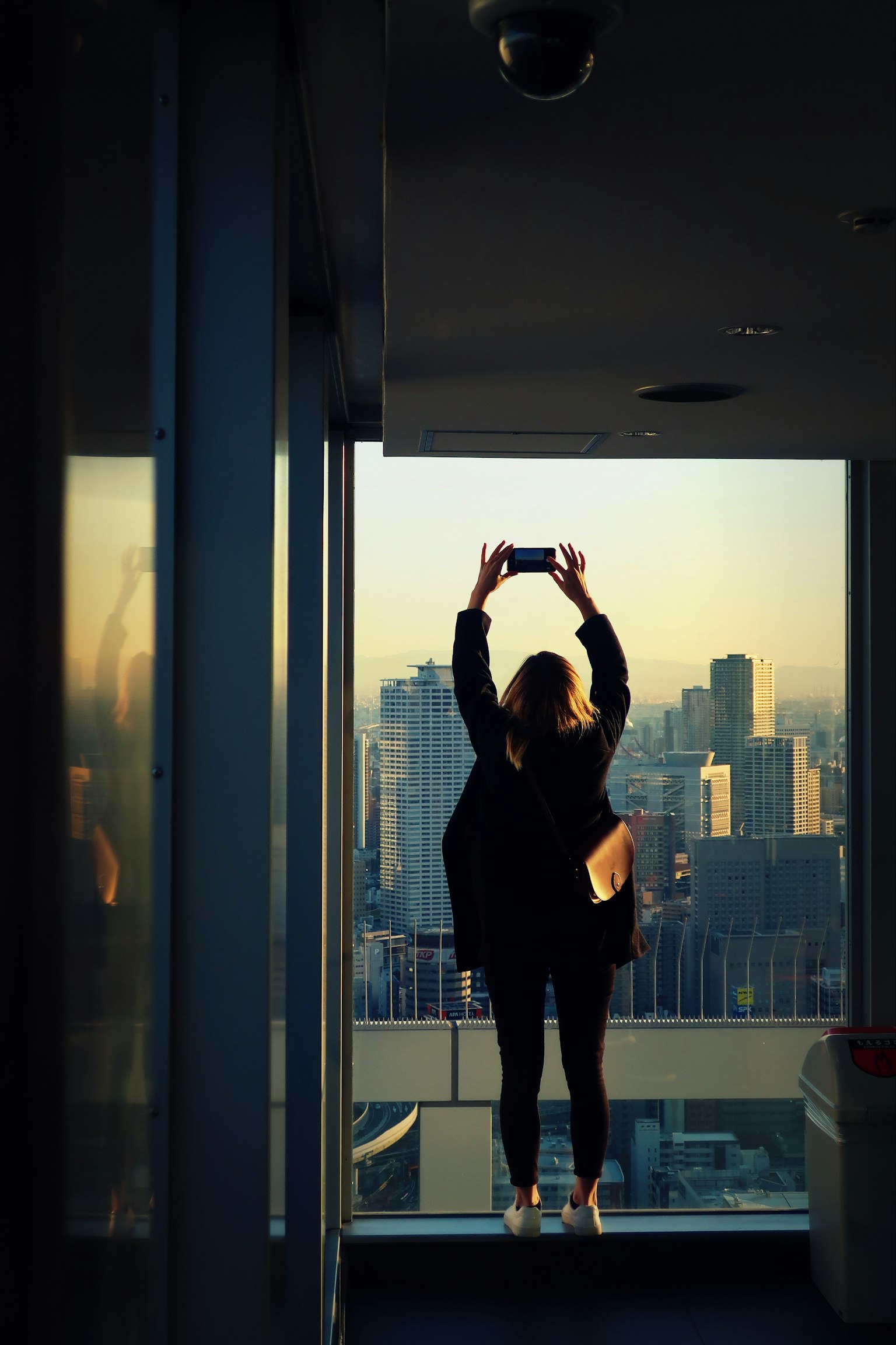 Silhouette d'une femme se tenant devant une fenêtre tenant un smartphone avec un paysage urbain en arrière-plan