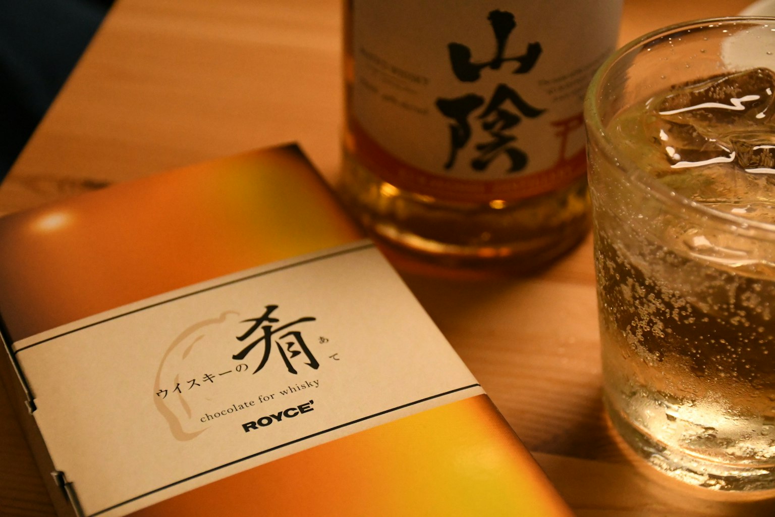 A package of fragrance next to a whiskey bottle and an ice-filled glass on a wooden table