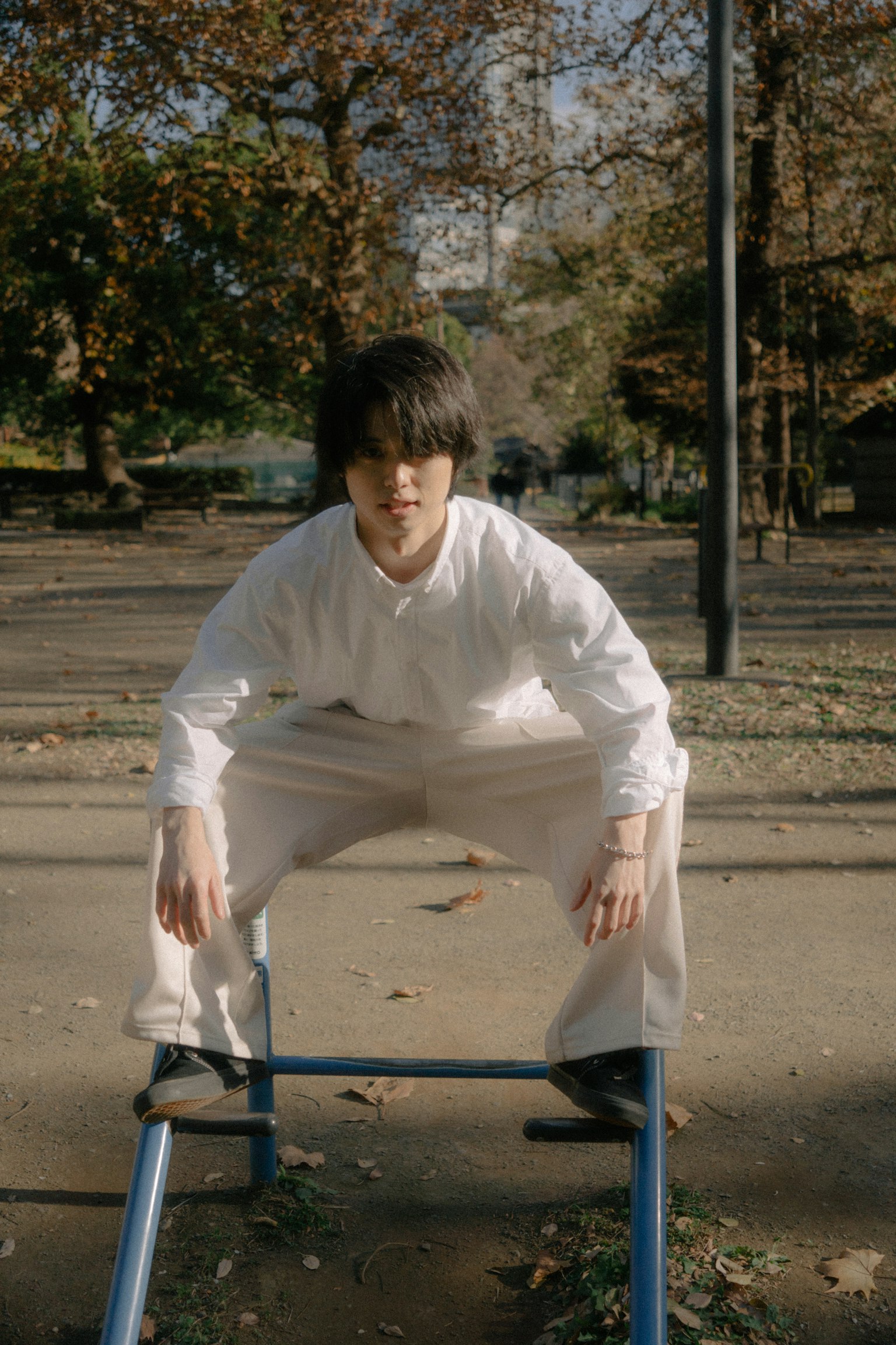 Young man wearing a white shirt and pants sitting on a blue apparatus in a park