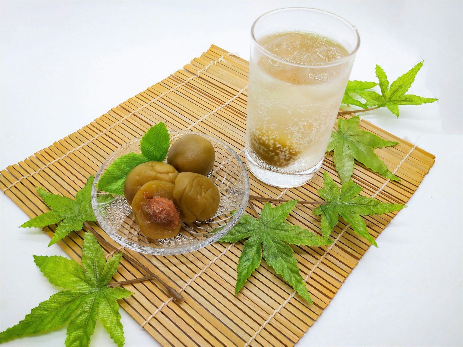 竹席上放著一杯飲料和一盤醃梅，周圍有綠色葉子