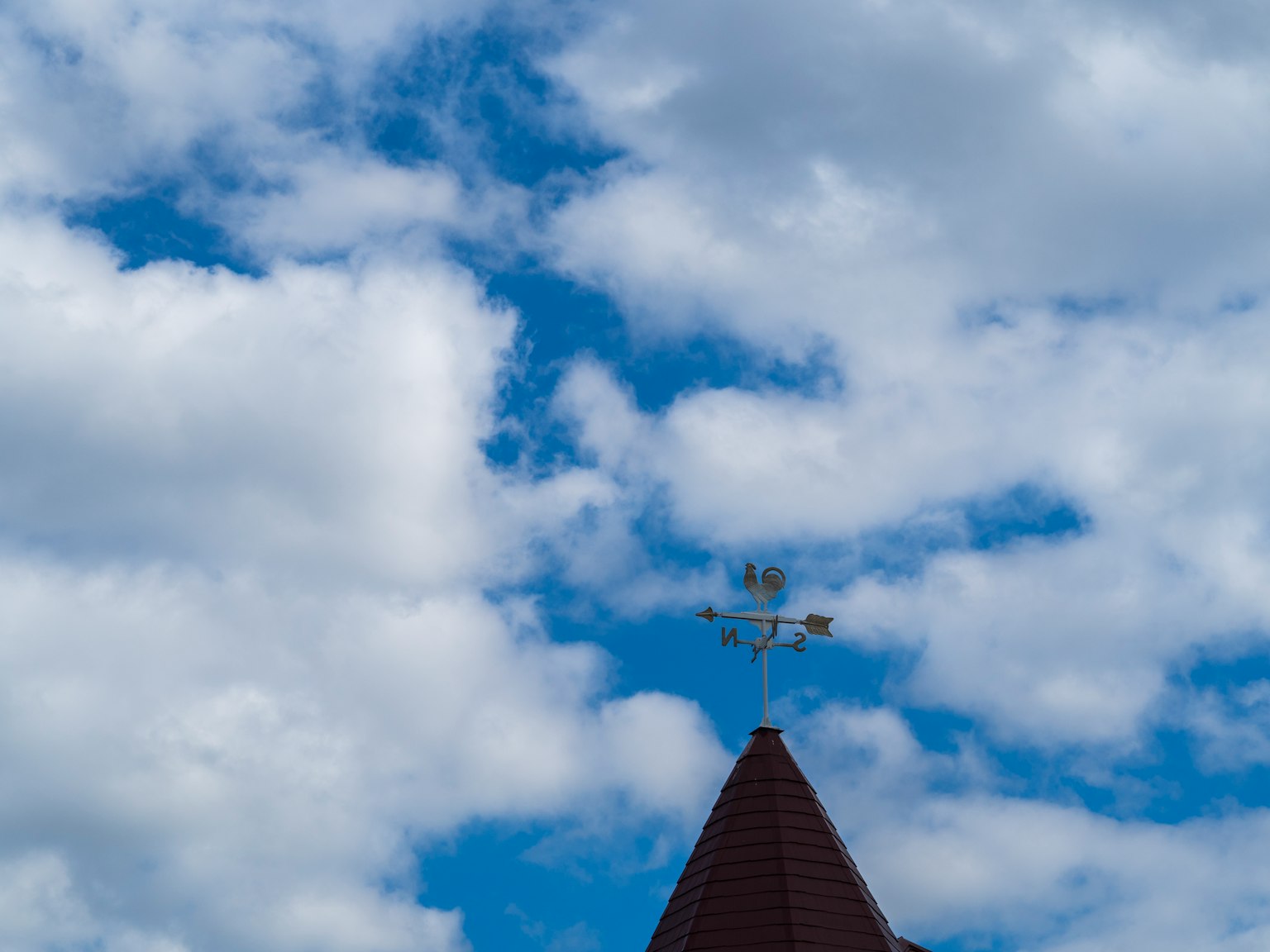 Blauer Himmel mit weißen Wolken und einem spitzen Dach mit einer Wetterfahne