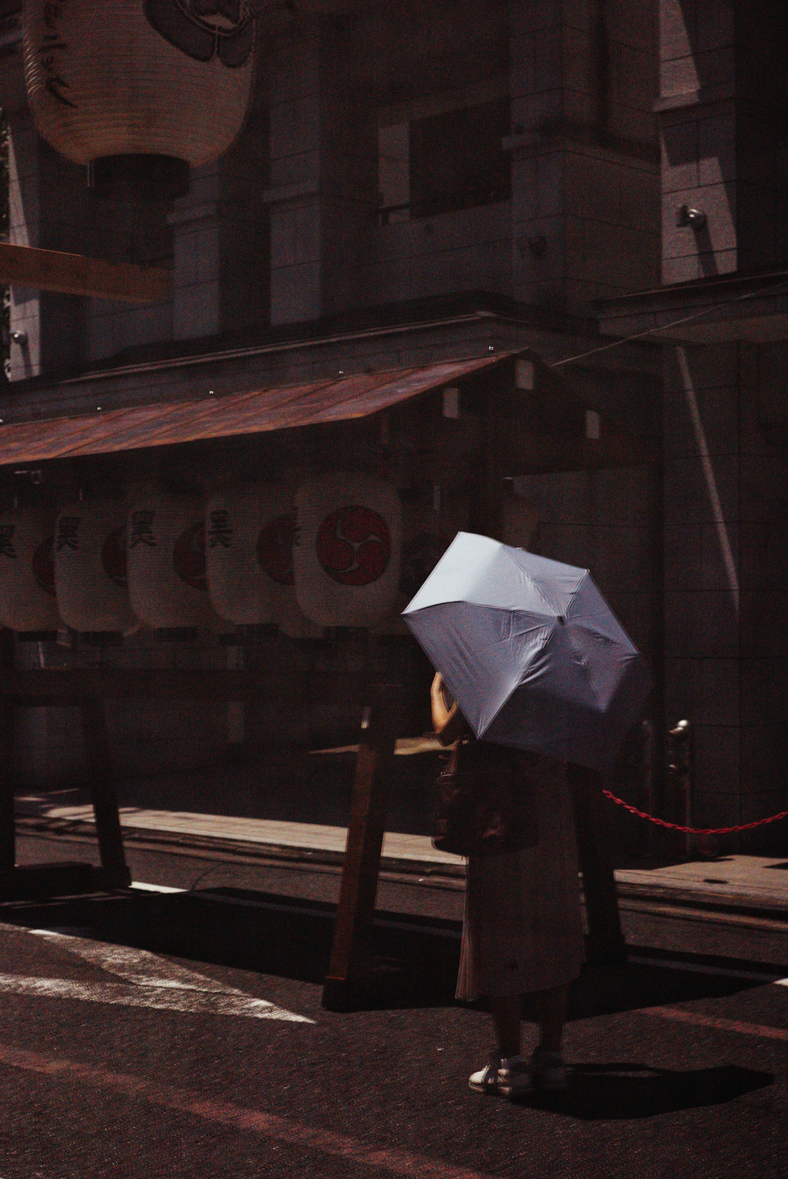 A person holding an umbrella walking in the street with a dark-toned background featuring traditional lanterns