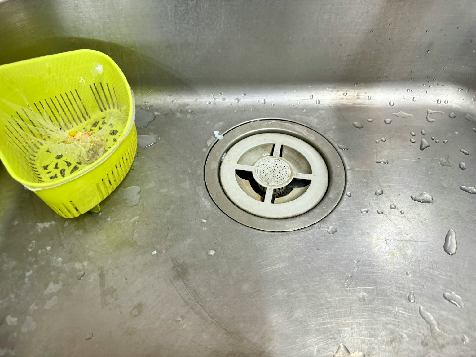 A view of a sink with water droplets a green strainer placed on the left side