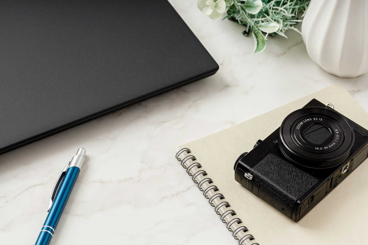Una macchina fotografica su un quaderno accanto a una penna blu e un laptop con una pianta sullo sfondo