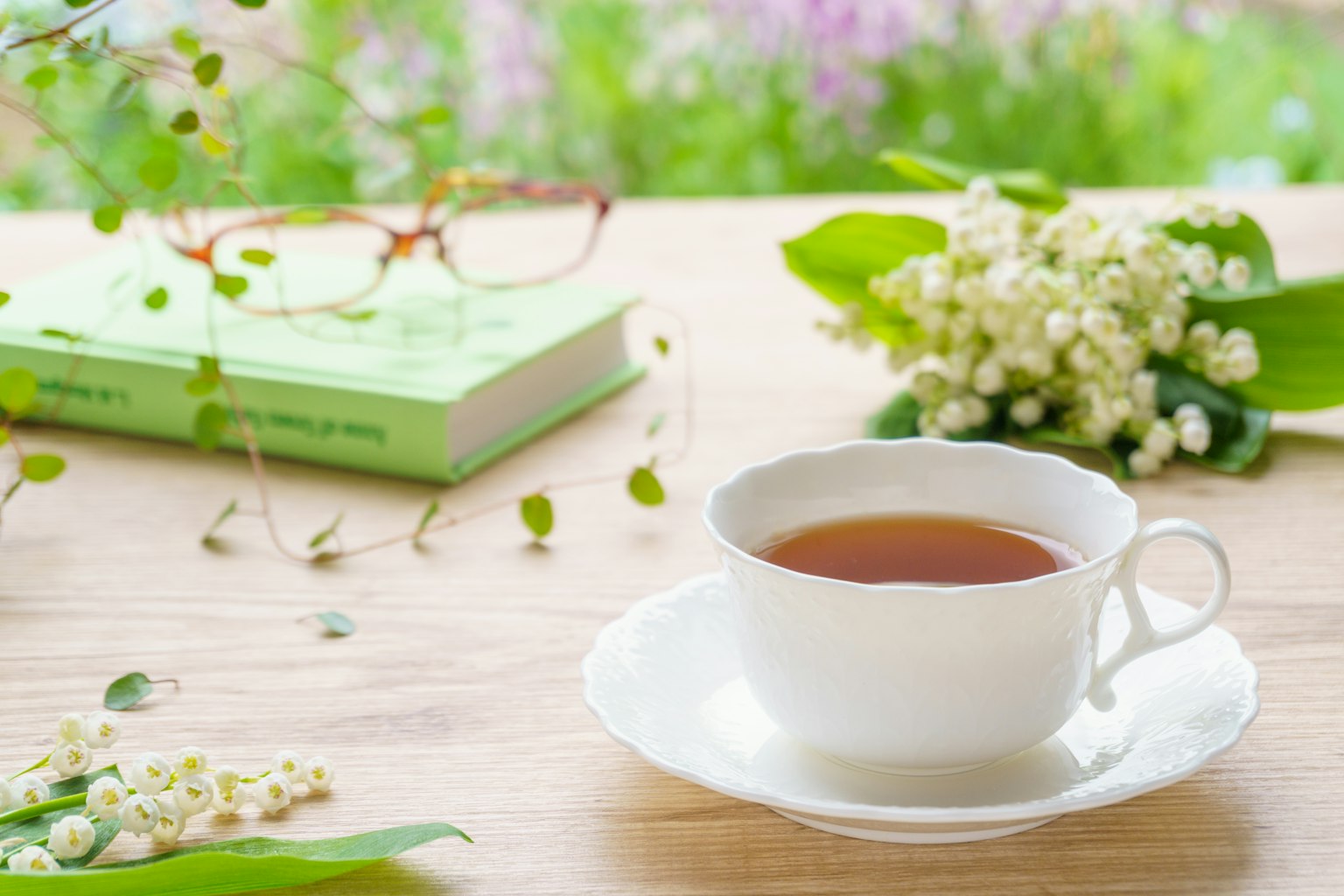 白色茶杯和茶托上放着茶旁边是绿色书籍眼镜和桌子上的花