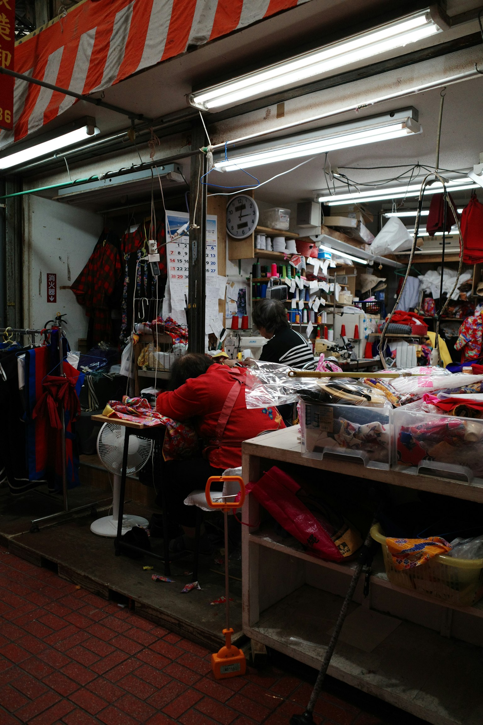 Un rincón de mercado estrecho con una persona vestida de rojo trabajando en una mesa rodeada de varias prendas y herramientas