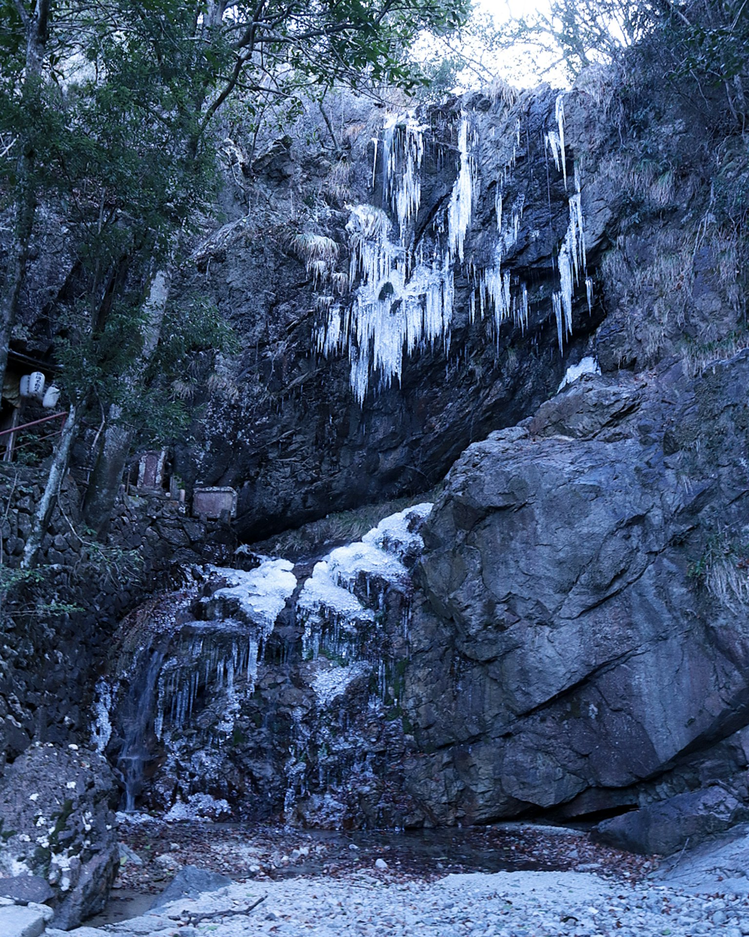 Cascata ghiacciata che scende da una scogliera rocciosa con stalattiti e ruscello ghiacciato