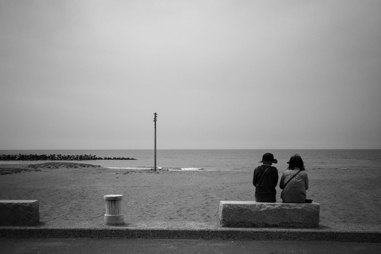 Dos personas sentadas en un banco junto a la playa con un mar tranquilo al fondo