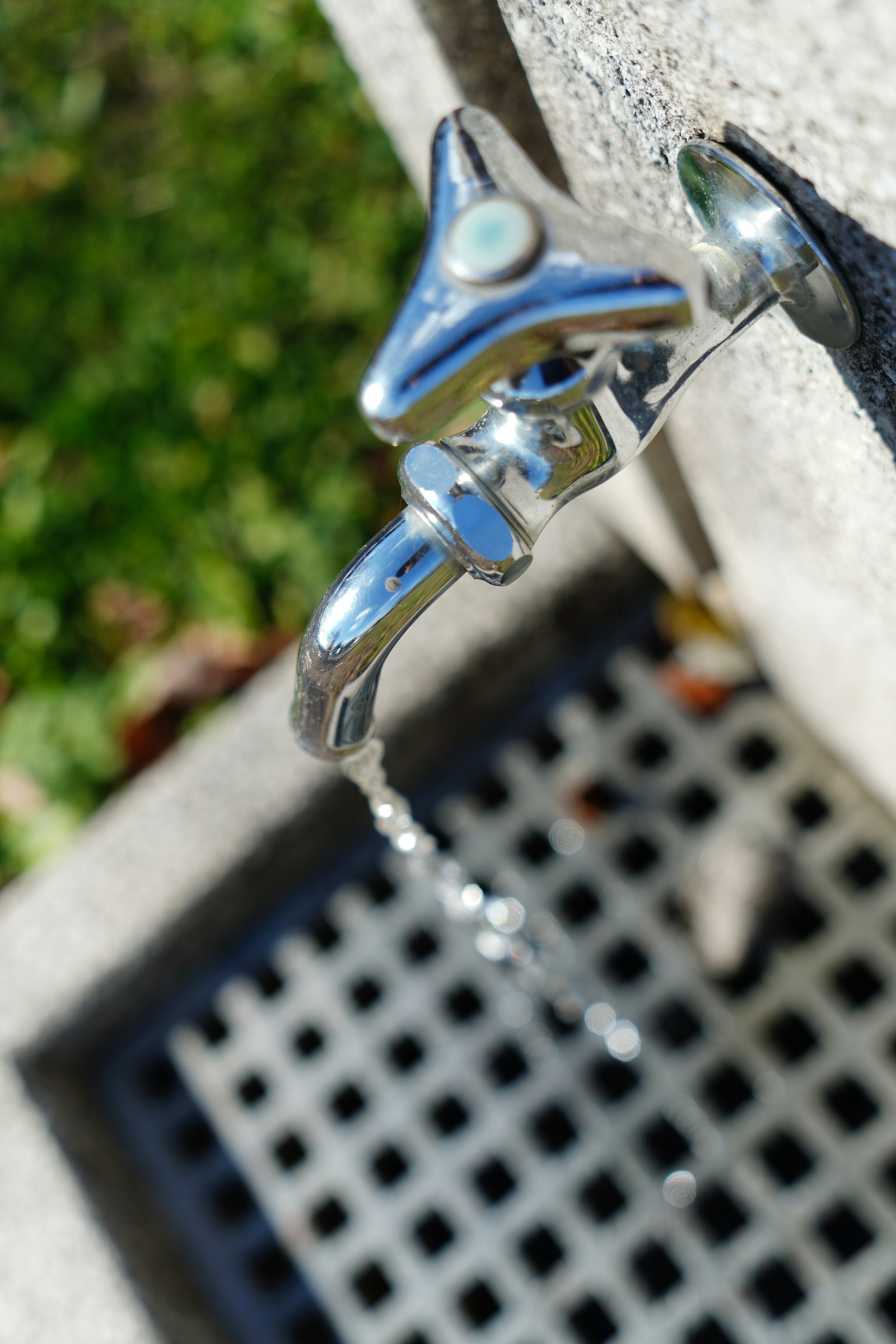 Photo of a faucet with water flowing from it The faucet is metallic and shiny with green grass in the background