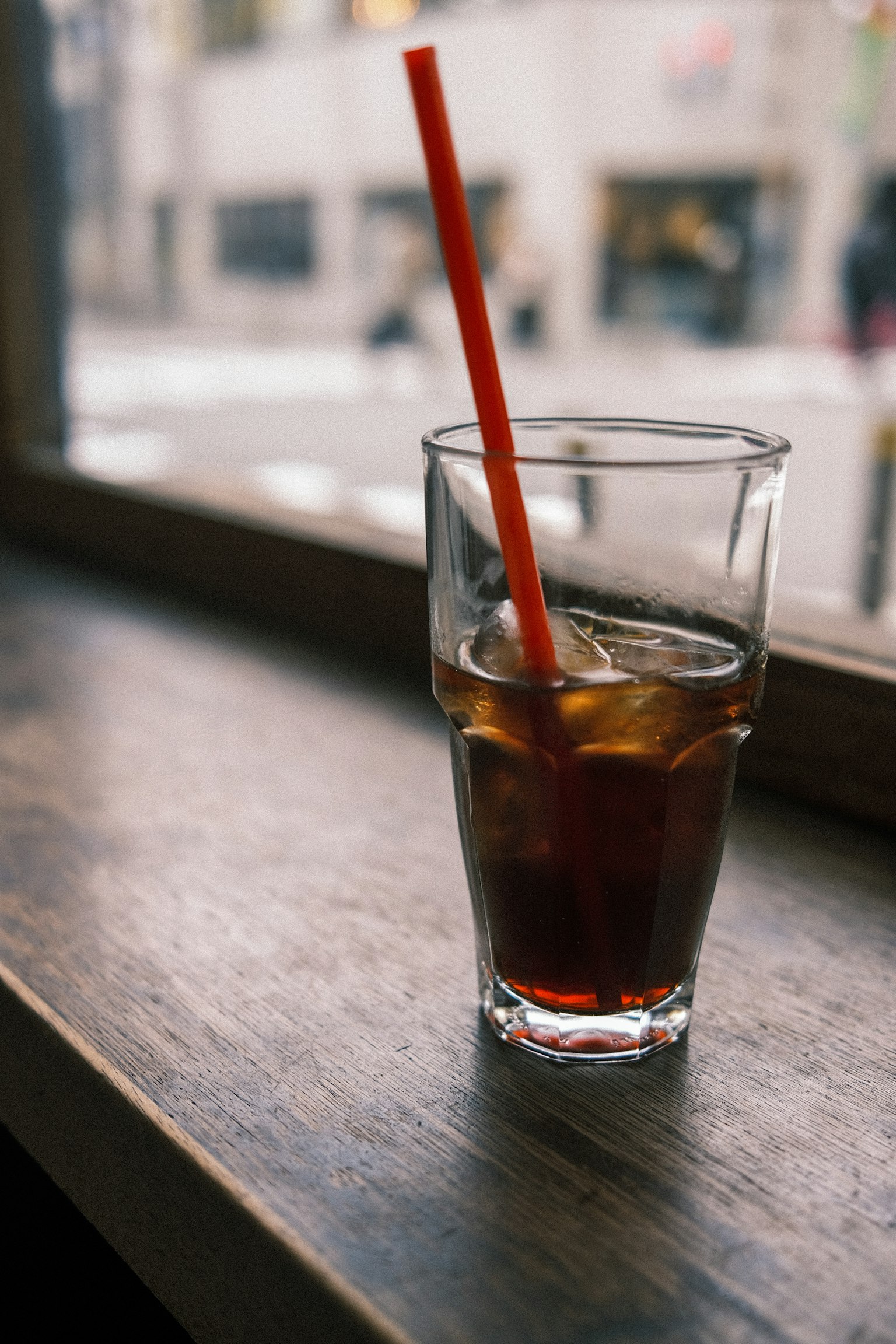 Un bicchiere di tè freddo con cubetti di ghiaccio e una cannuccia rossa posato vicino a una finestra