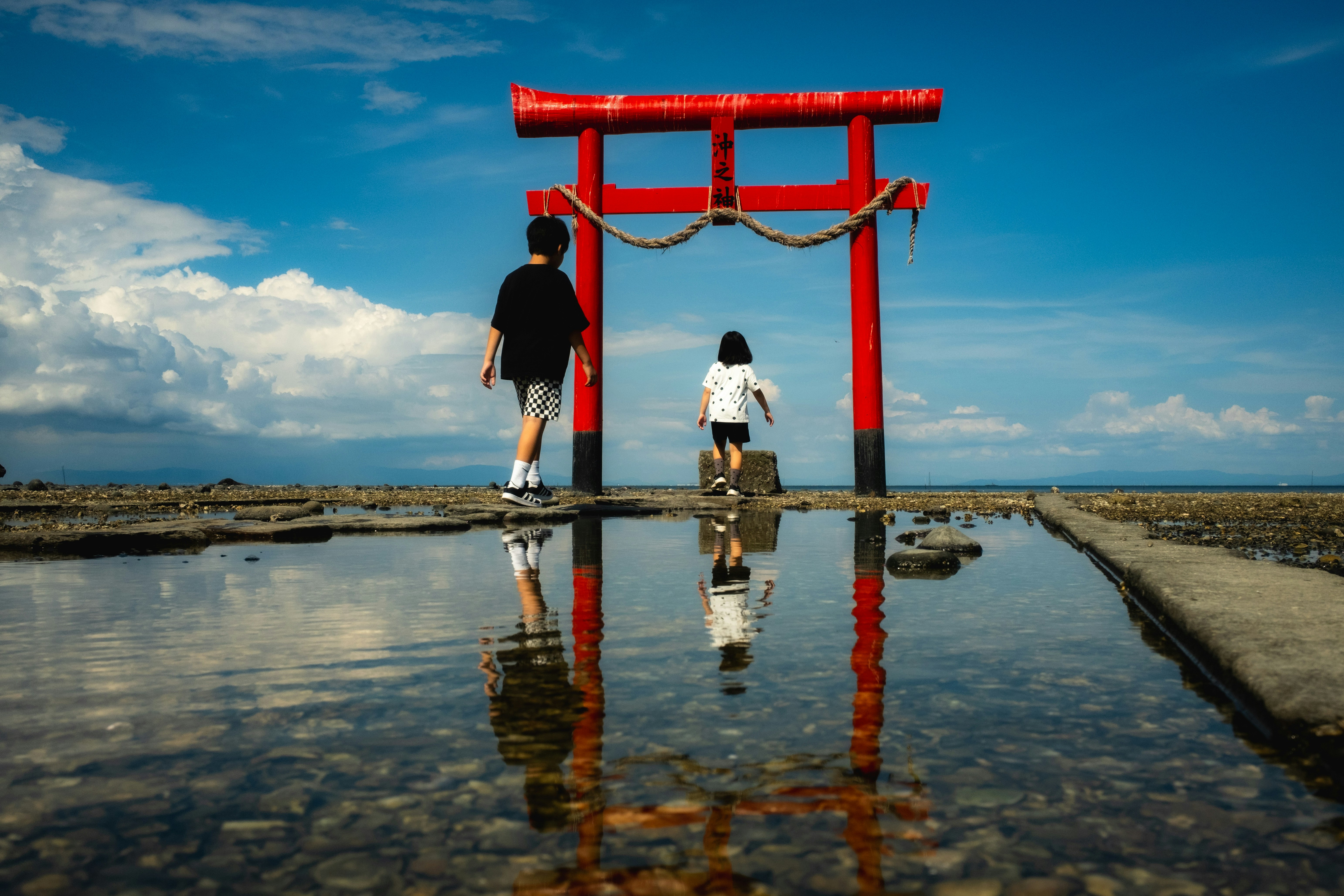 赤い鳥居の前に立つ子供たちのシルエットと青い空の反射