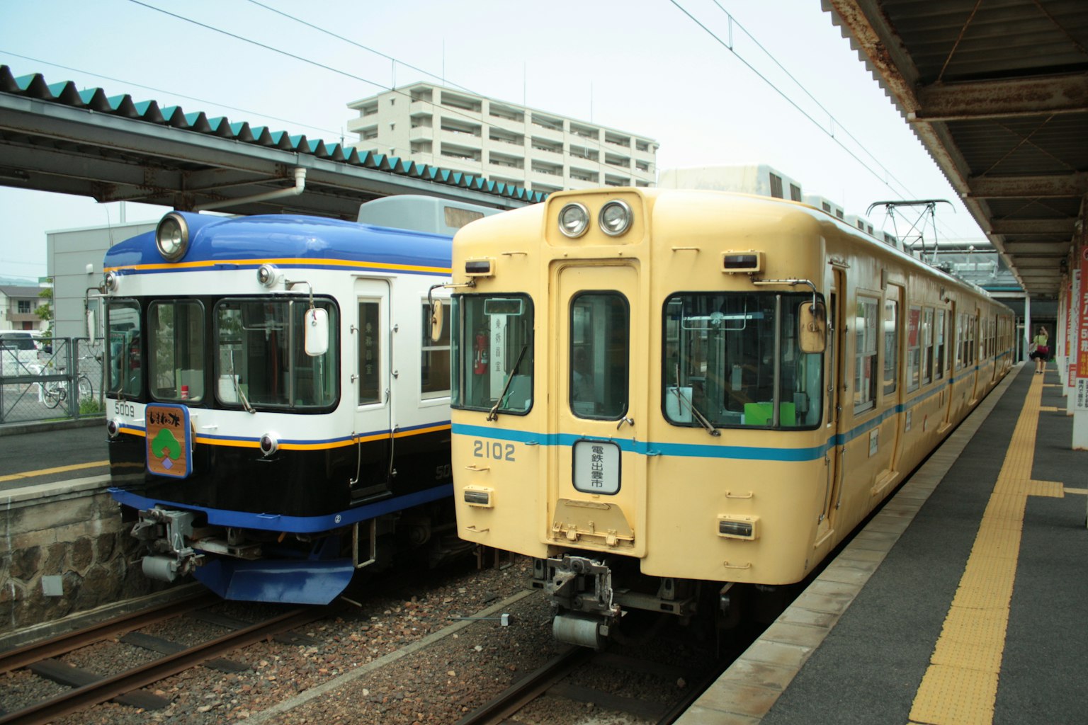 Dua kereta diparkir berdampingan di stasiun dengan kereta biru dan kereta kuning